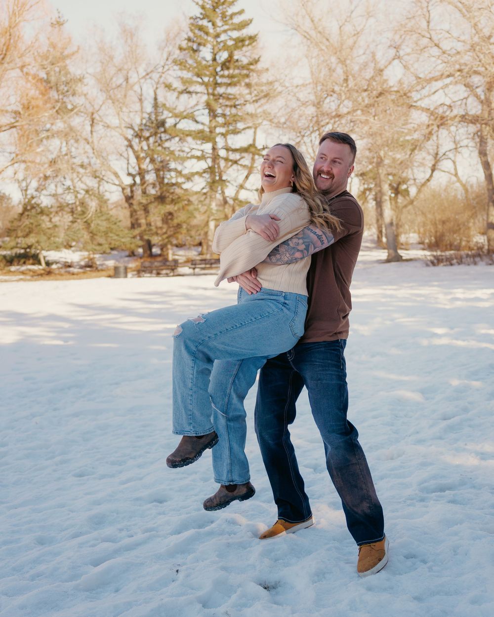 Joyful couple laughing as the man lifts the woman in a sunny snow-covered winter landscape.