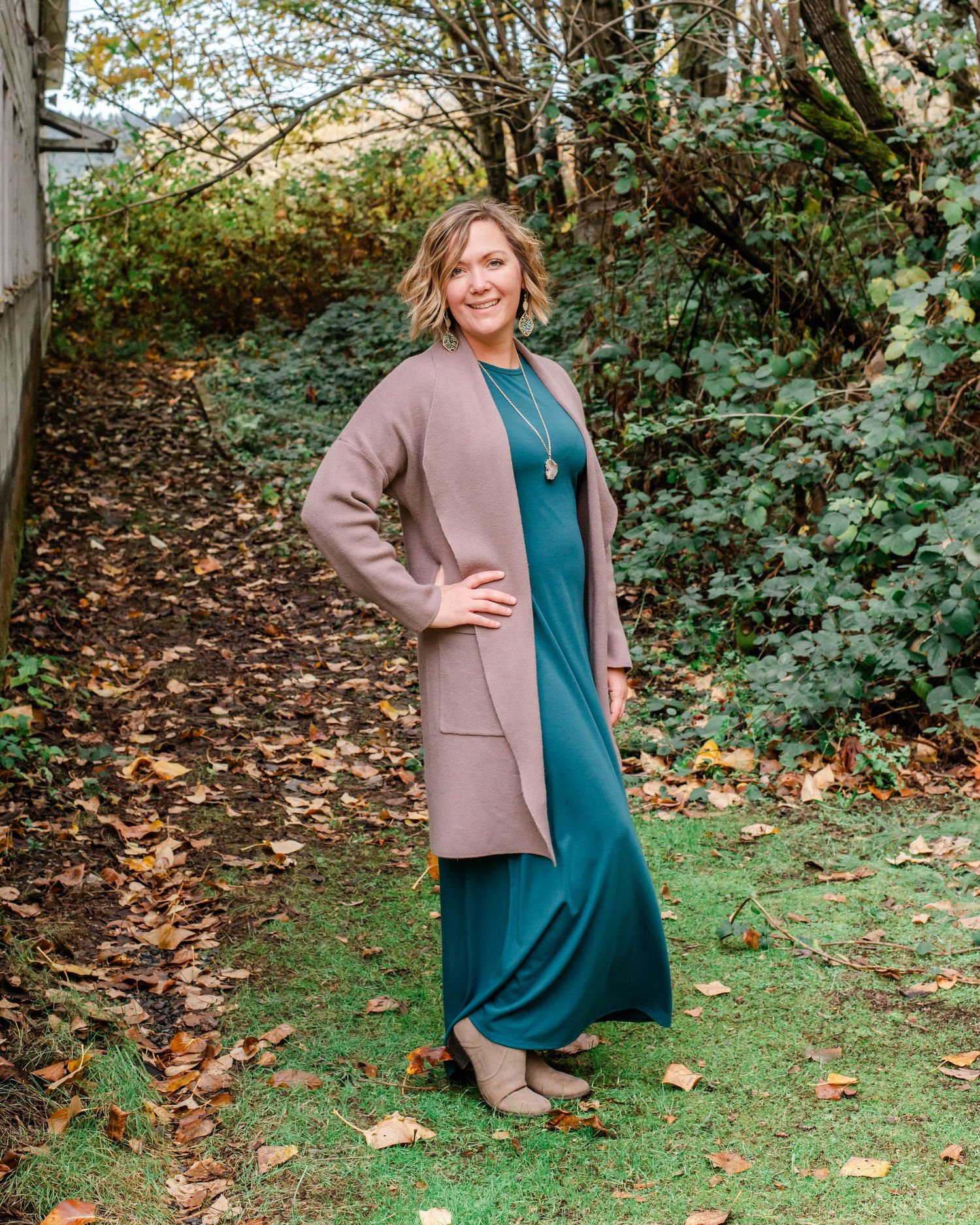 Woman wearing teal dress and brown cardigan stands in autumn garden setting.