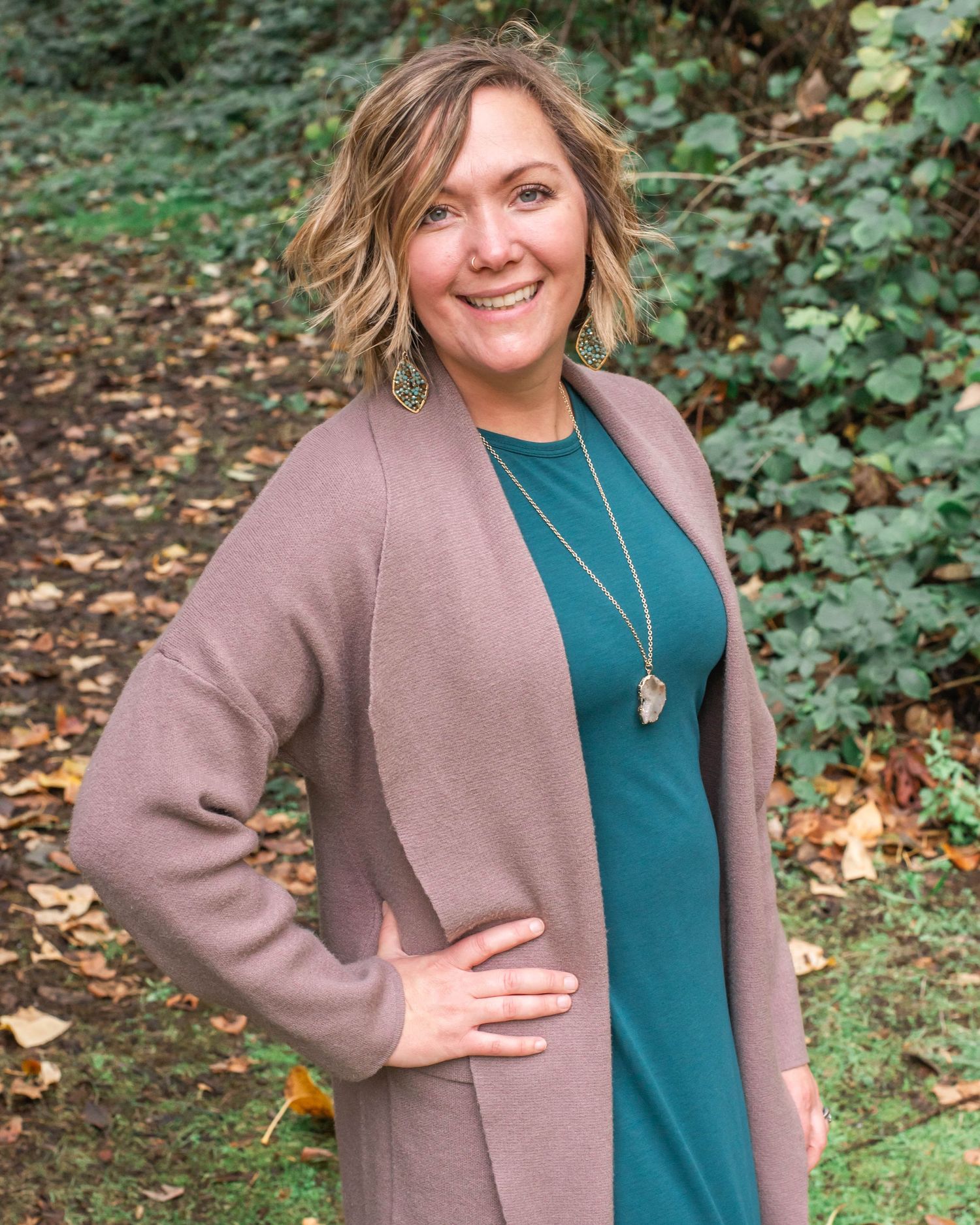 Woman in teal dress and mauve cardigan poses outdoors against green foliage.