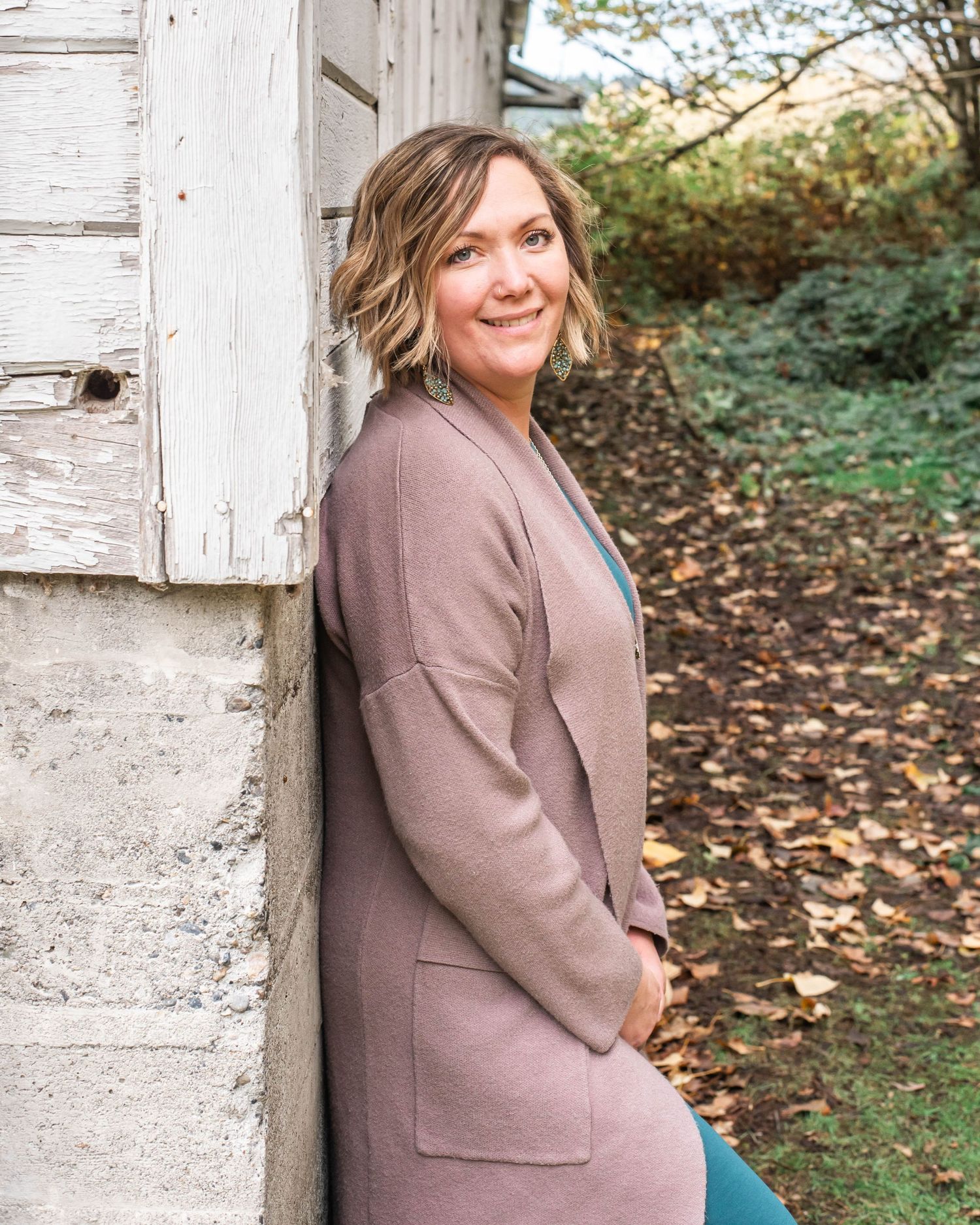 Woman in teal dress and brown blazer poses against white rustic wall outdoors.