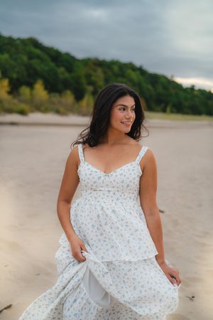 Woman in white floral sundress sits gracefully on beach with cloudy sky backdrop.
