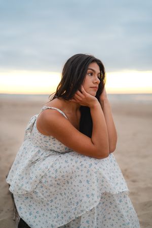 Woman in floral dress poses thoughtfully on sandy beach at sunset.