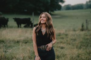 Person in black outfit standing in a grassy field with cattle in the background.