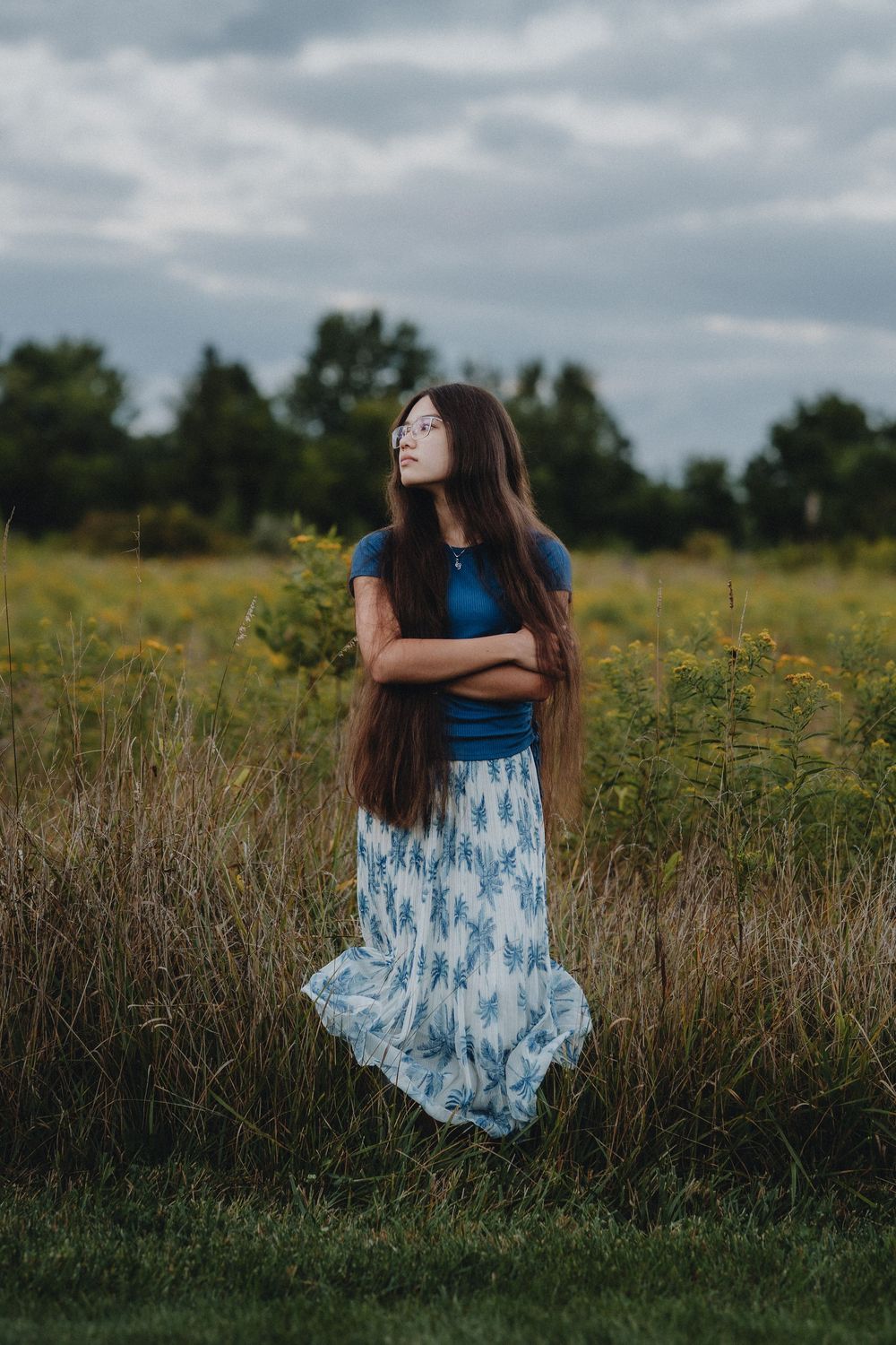 Figure in blue dress twirling in tall grass field under cloudy skies.