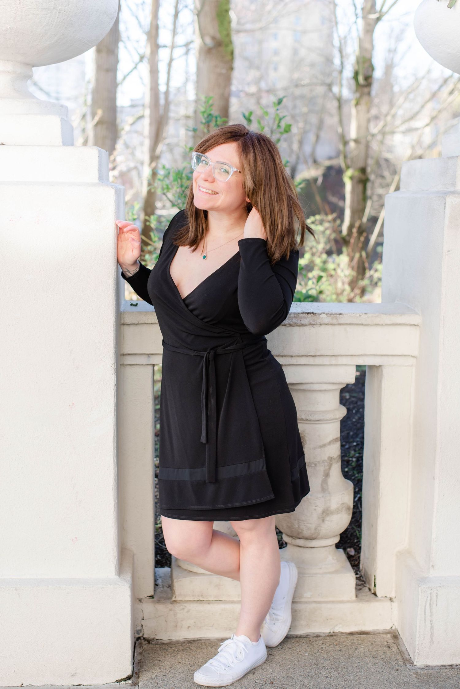 Person in black top poses playfully on stone ledge outdoors with tree branches in background.