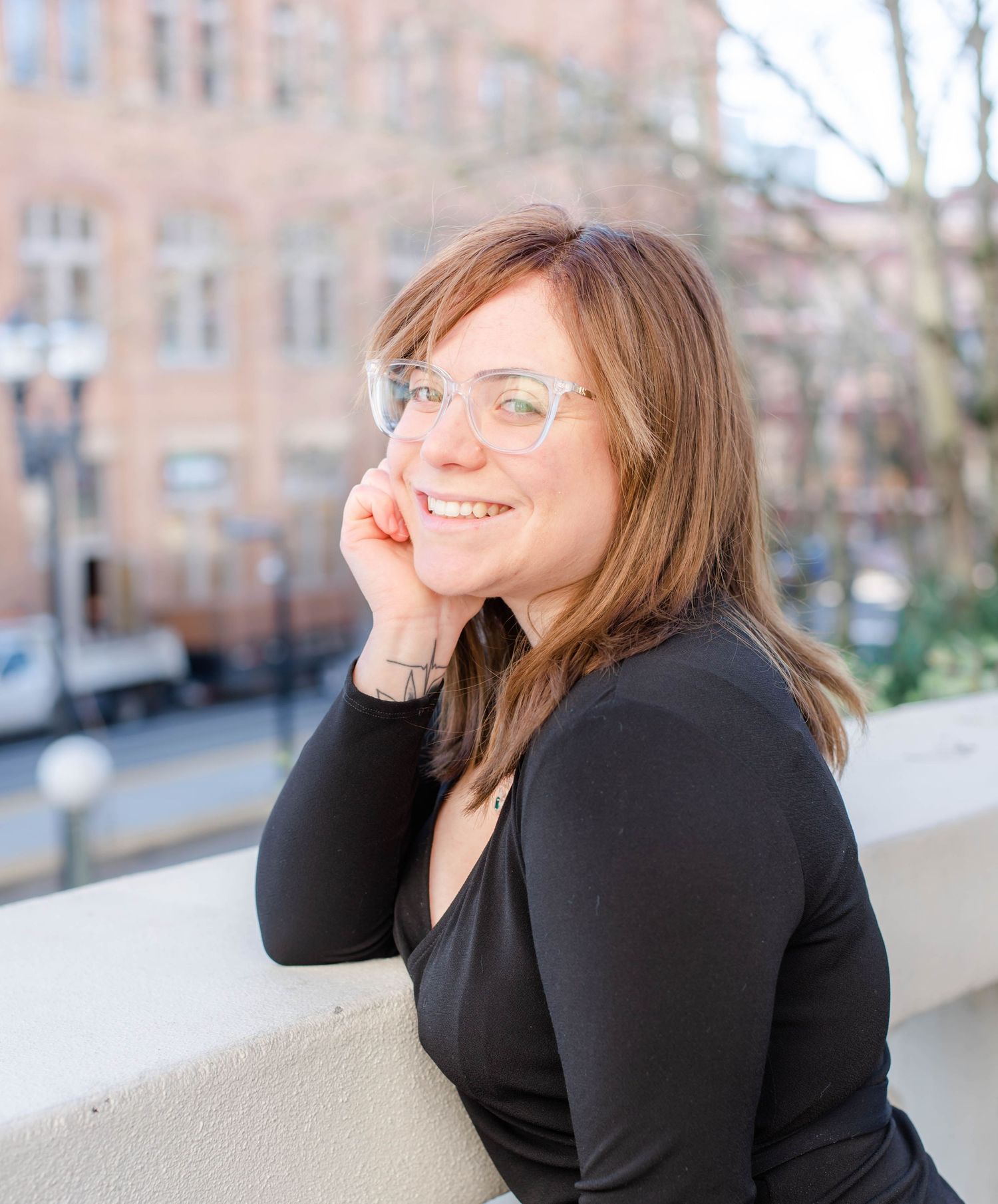 Person in black top lounges on stone wall smiling against blurred city background.