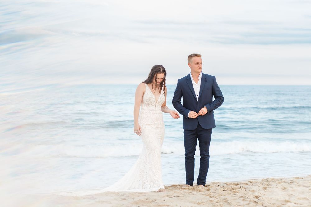 A couple in wedding attire walks along a scenic beach with waves and cloudy skies in the background.