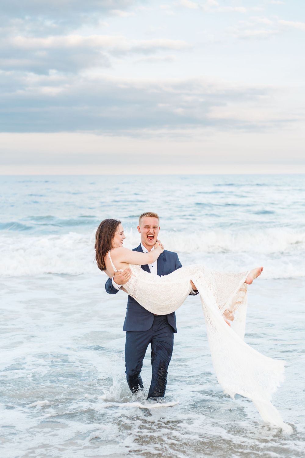 A playful wedding couple splashing in ocean waves as the groom carries the bride.