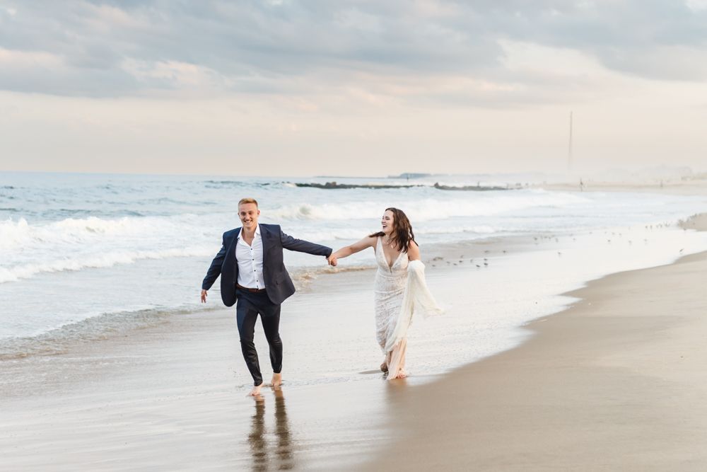 A couple runs playfully together along a misty beach shore at sunset with crashing waves in the background.