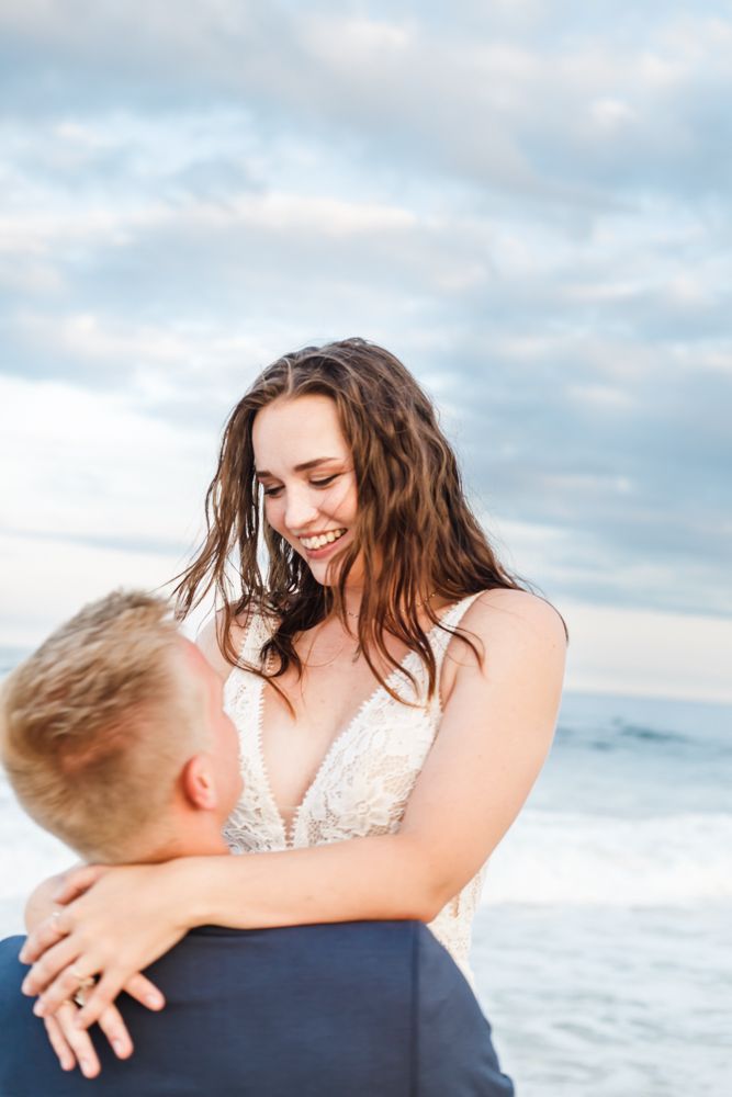 Romantic beach photoshoot at sunset with couple embracing by the ocean waves and cloudy sky.
