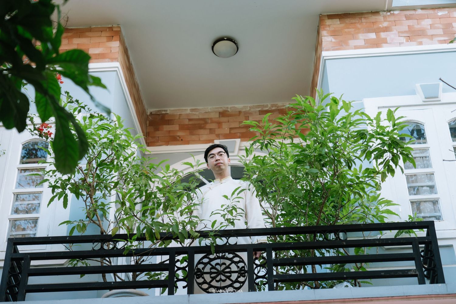 Young Vietnamese man in white ao dai standing on balcony surrounded by lush green plants.