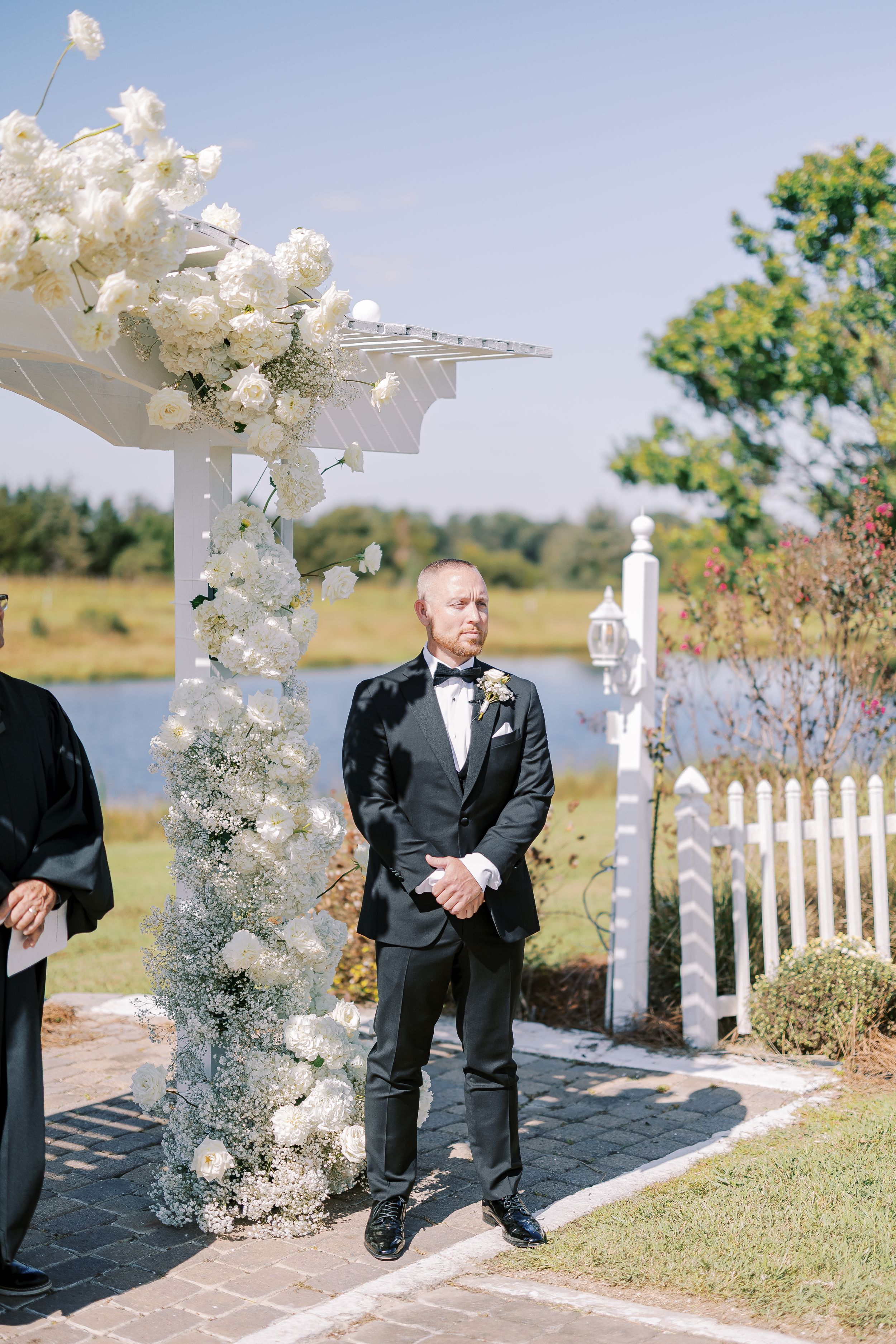 A series of elegant white floral wedding arch arrangements against a waterfront backdrop on a sunny day.