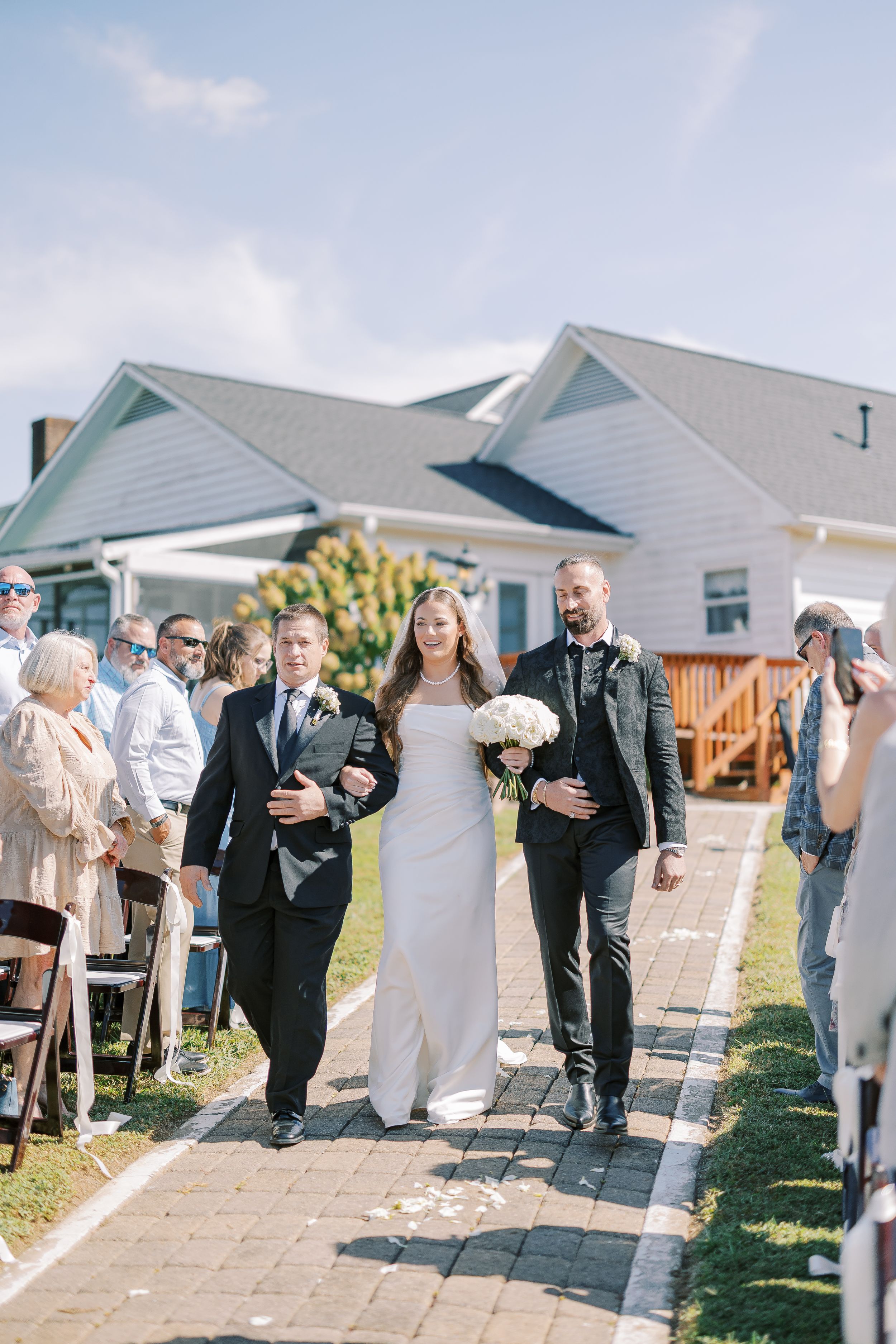 Wedding guests stand together outdoors along a paved walkway during an outdoor ceremony celebration.