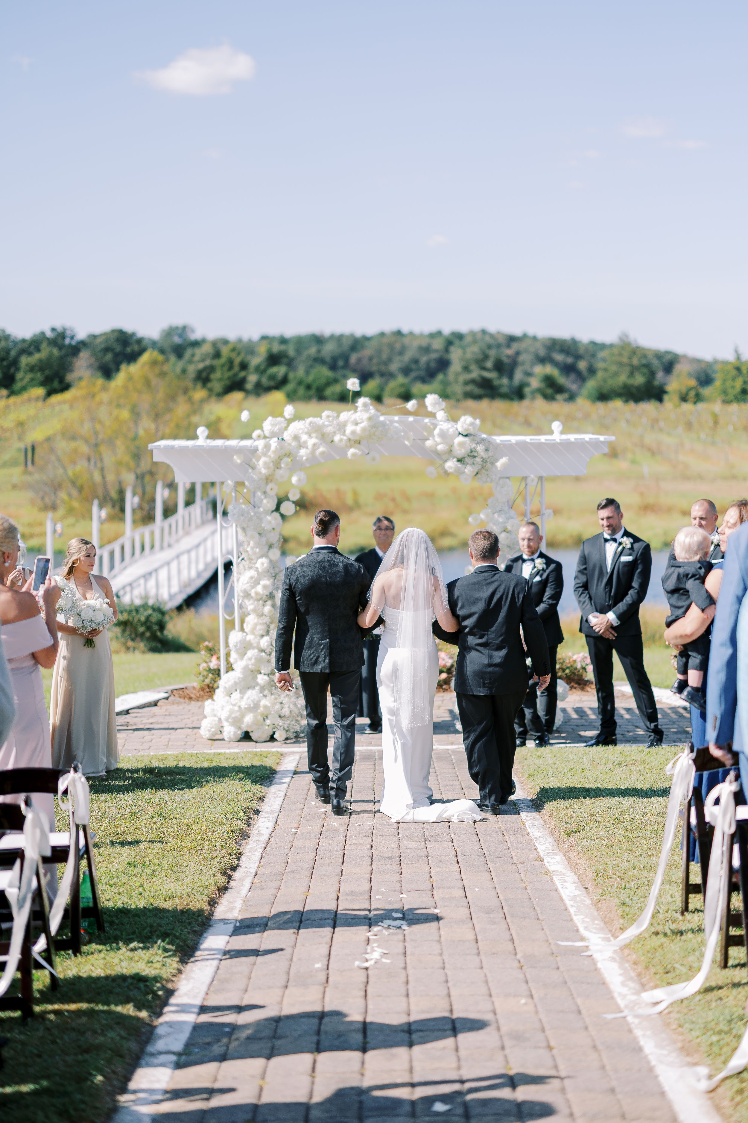 Wedding party walking down outdoor pathway lined with white chairs and floral decorations on a sunny day.