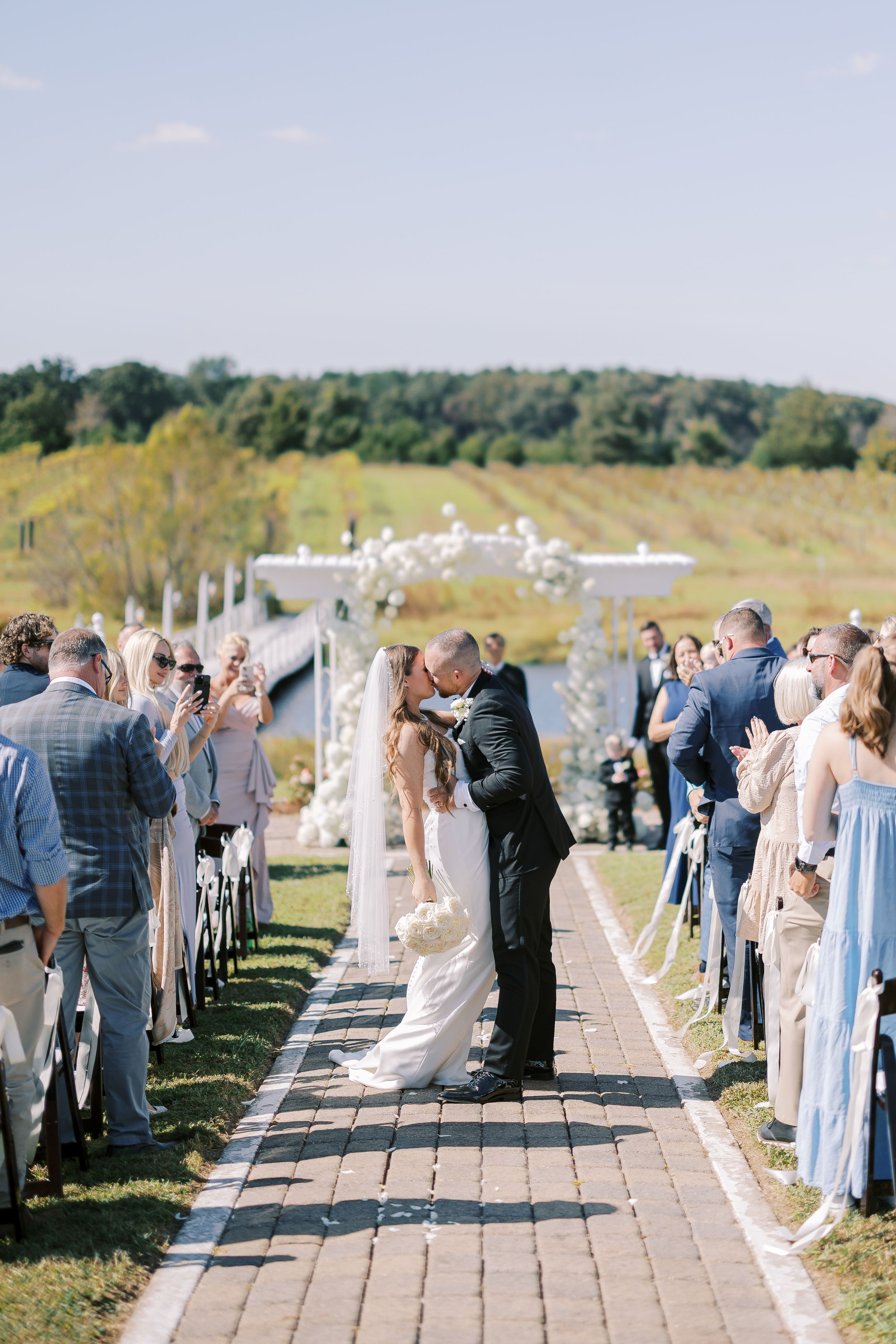 Wedding guests line both sides of a scenic waterfront path during an outdoor ceremony celebration.