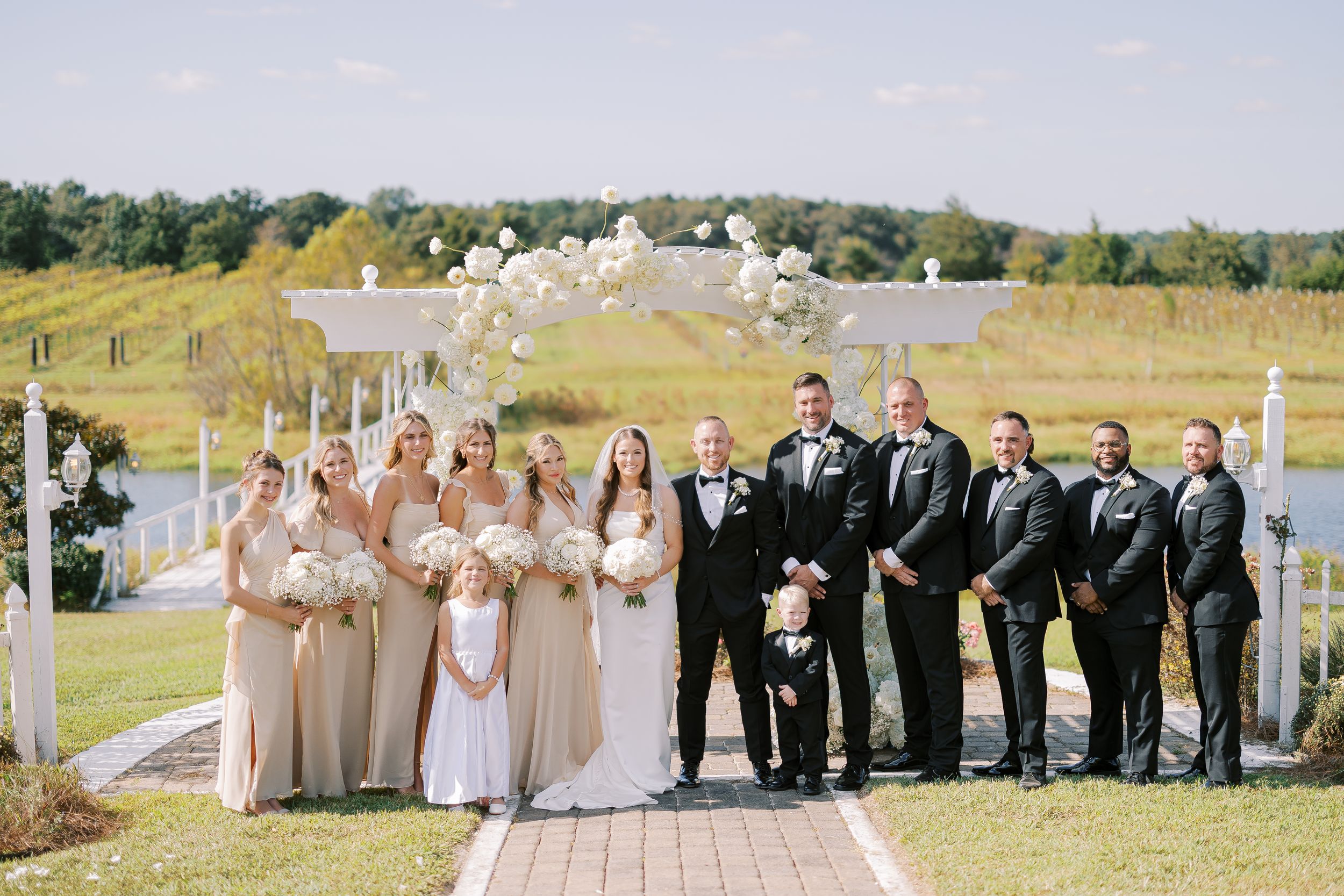 Wedding party poses elegantly on a grassy hillside with luxurious white floral arch decorations and natural landscaping.