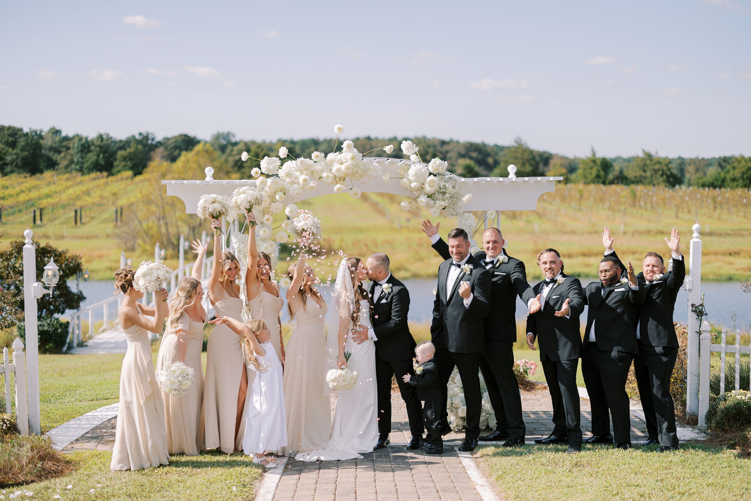 Wedding party in pink and black formal attire poses together outdoors with scenic landscape in background.