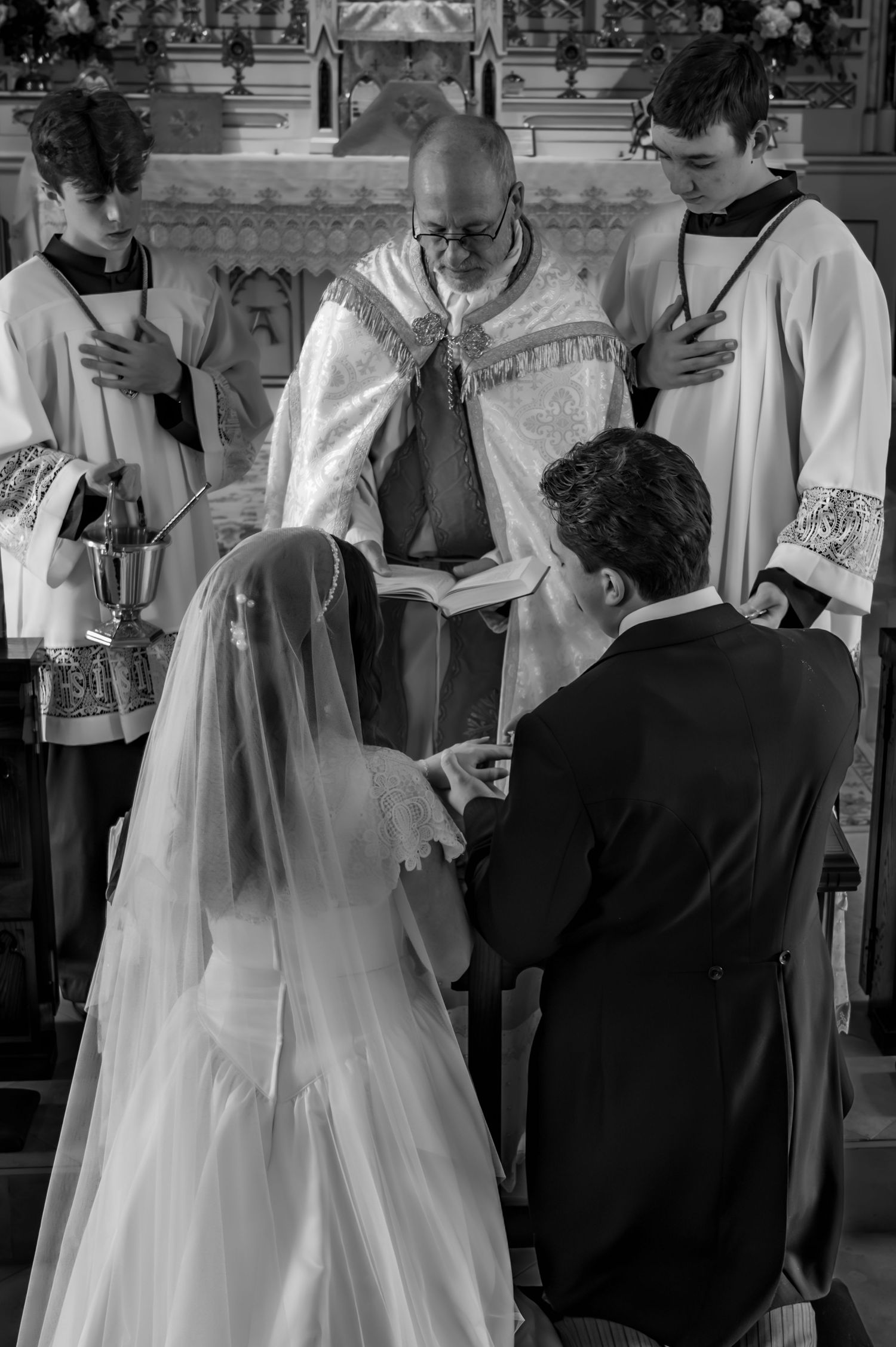 Black and white photo of bride and groom exchanging vows before a priest at a church altar.