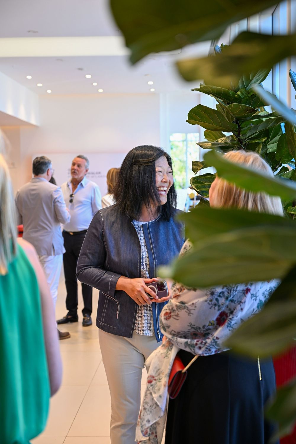 Business professionals socialize during an office gathering in a bright modern lobby area near indoor plants.