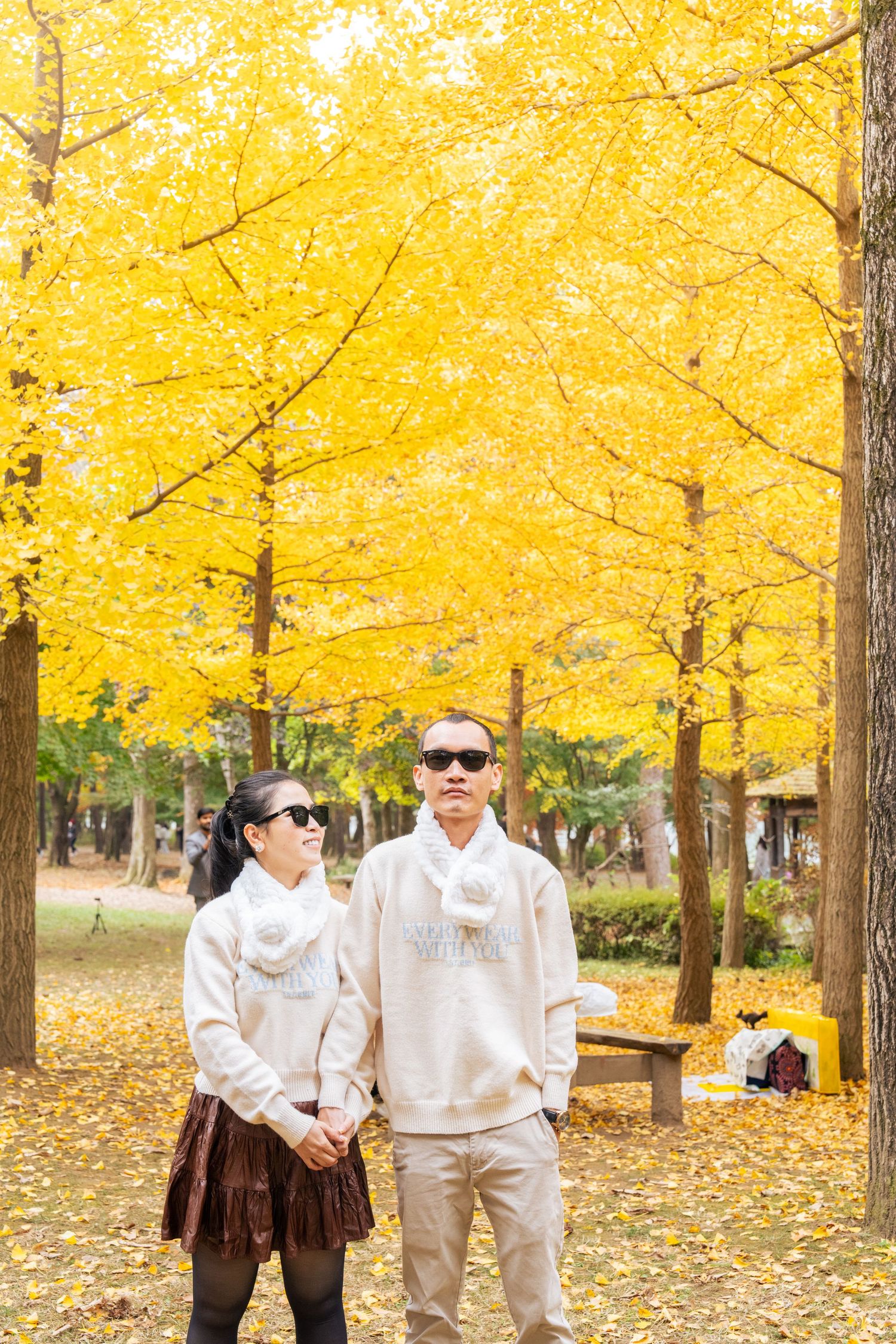 Group of people walking beneath vibrant yellow ginkgo trees in an autumn park with fallen leaves on the ground.