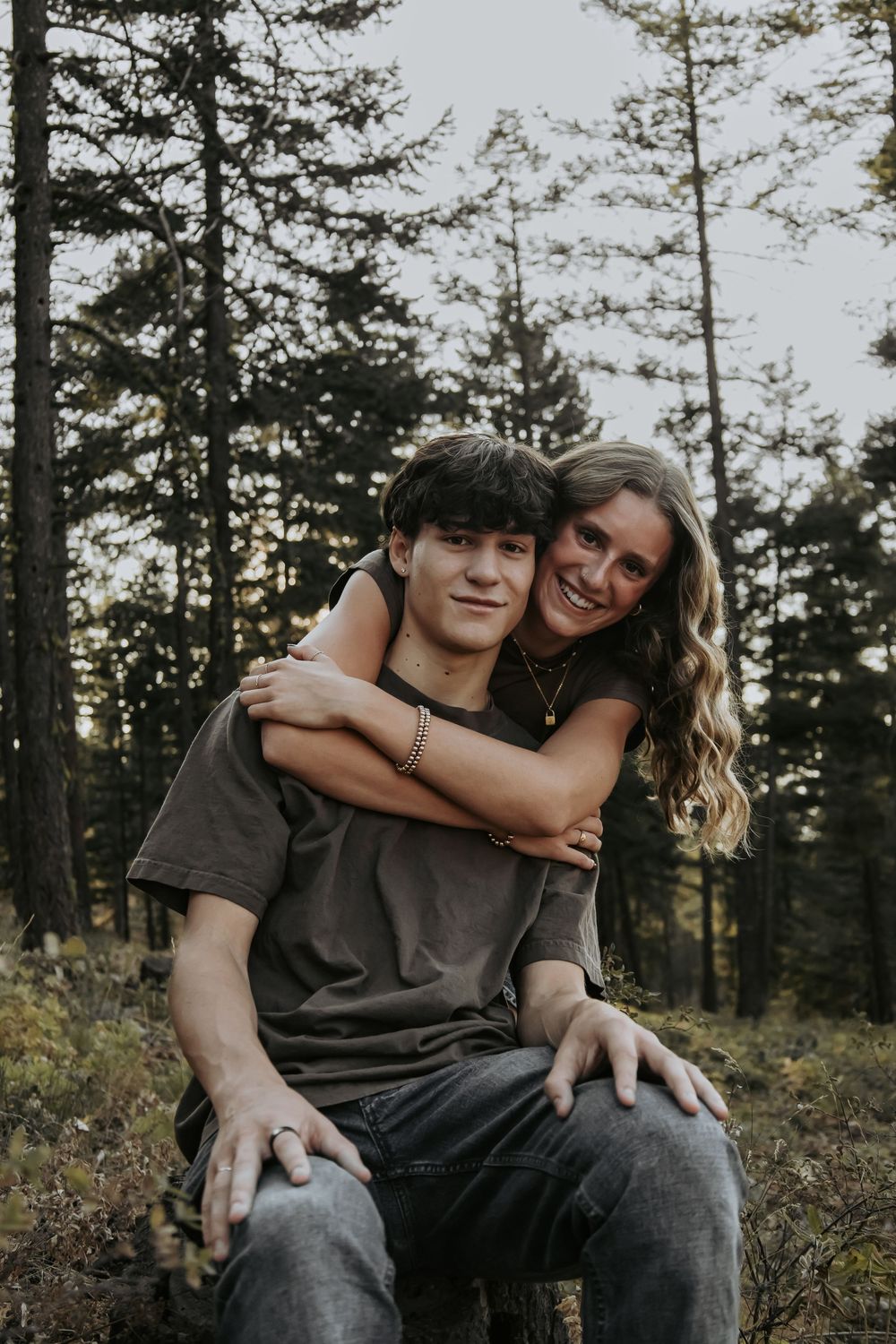 Couple sharing affectionate moment among tall pine trees at dusk.