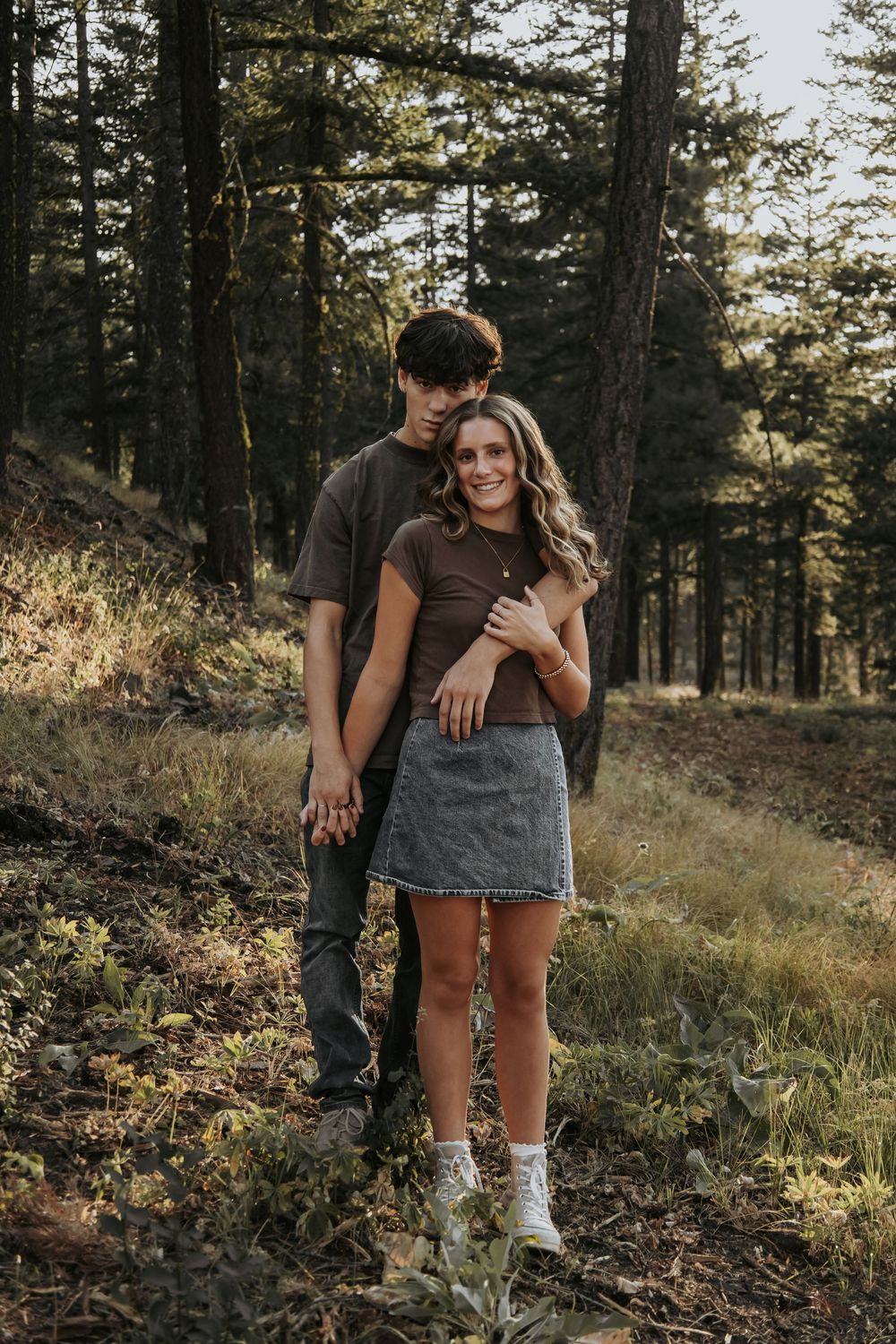 A couple poses playfully among pine trees on a hillside trail.