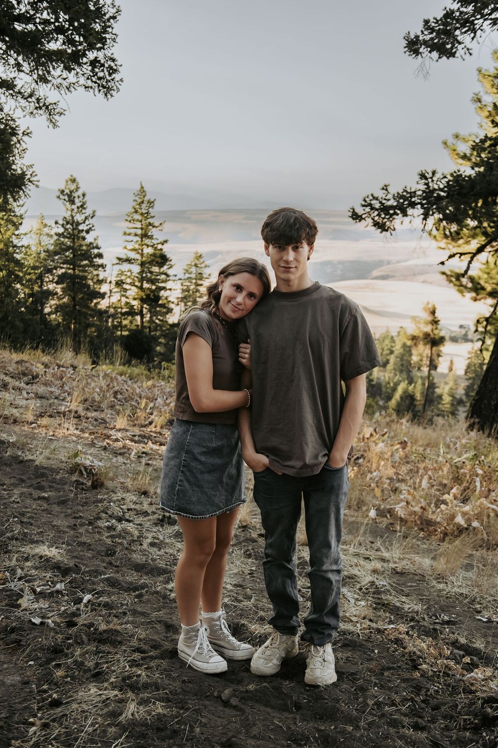 A couple stands close together on a mountainside with evergreen trees in the background.