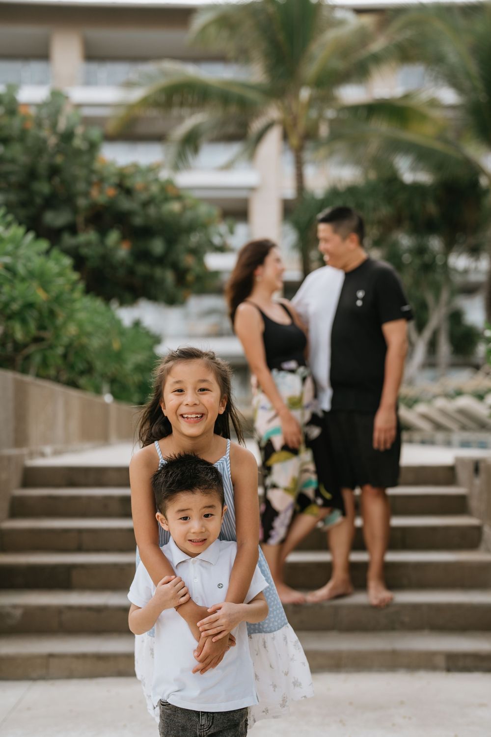 A family plays together at a tropical resort with palm trees and stone steps in the background.
