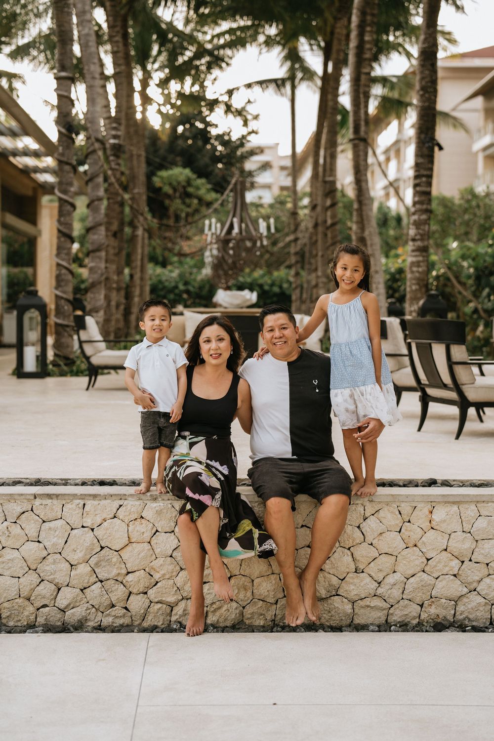 Family members sit casually on a stone wall surrounded by palm trees at a resort.