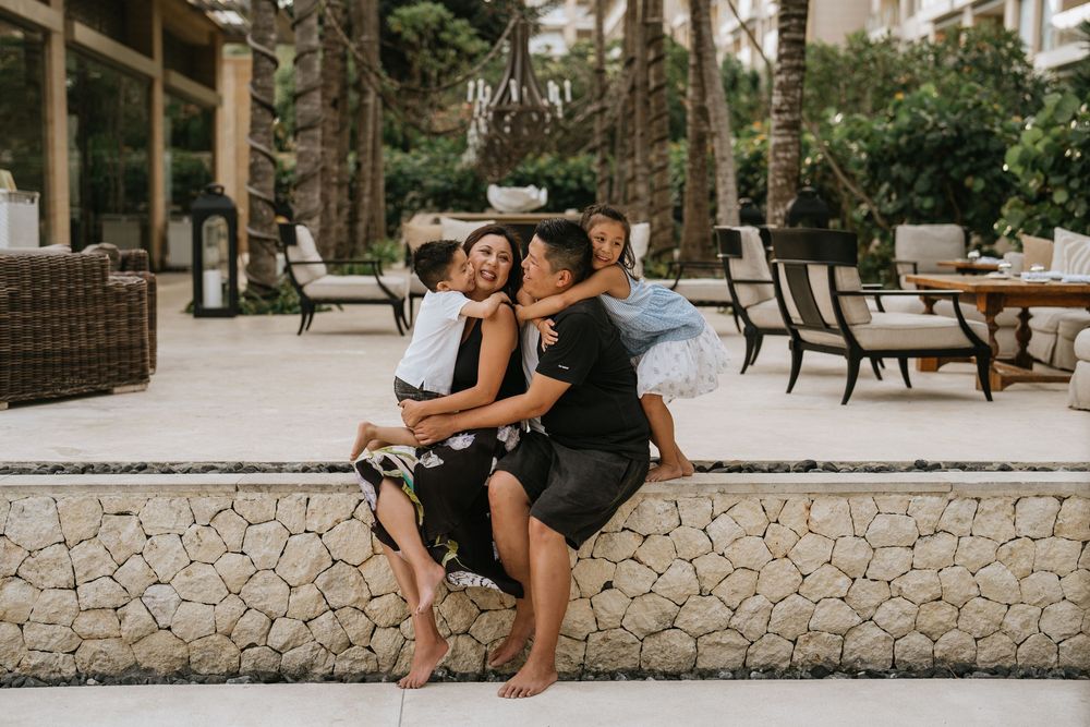 Family members share intimate moments on a stone wall at a tropical resort.