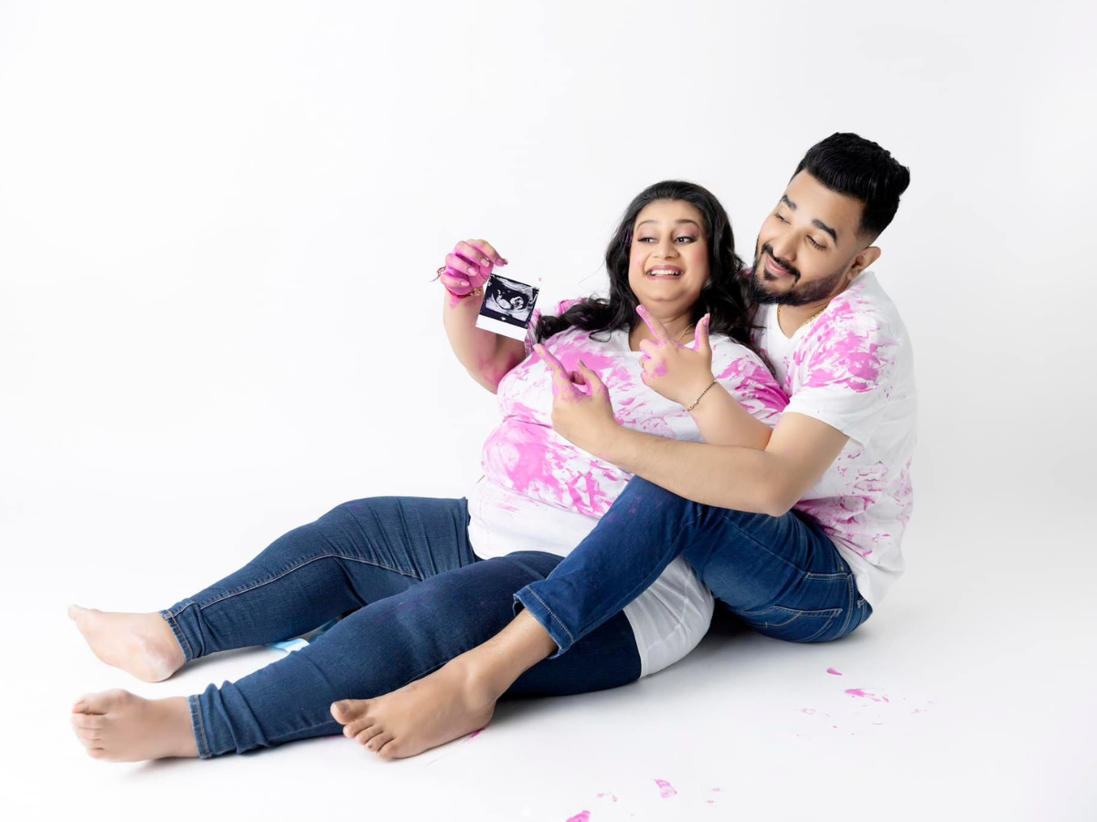 Two people in white shirts with pink paint stains sit and smile together while holding a camera on white backdrop.