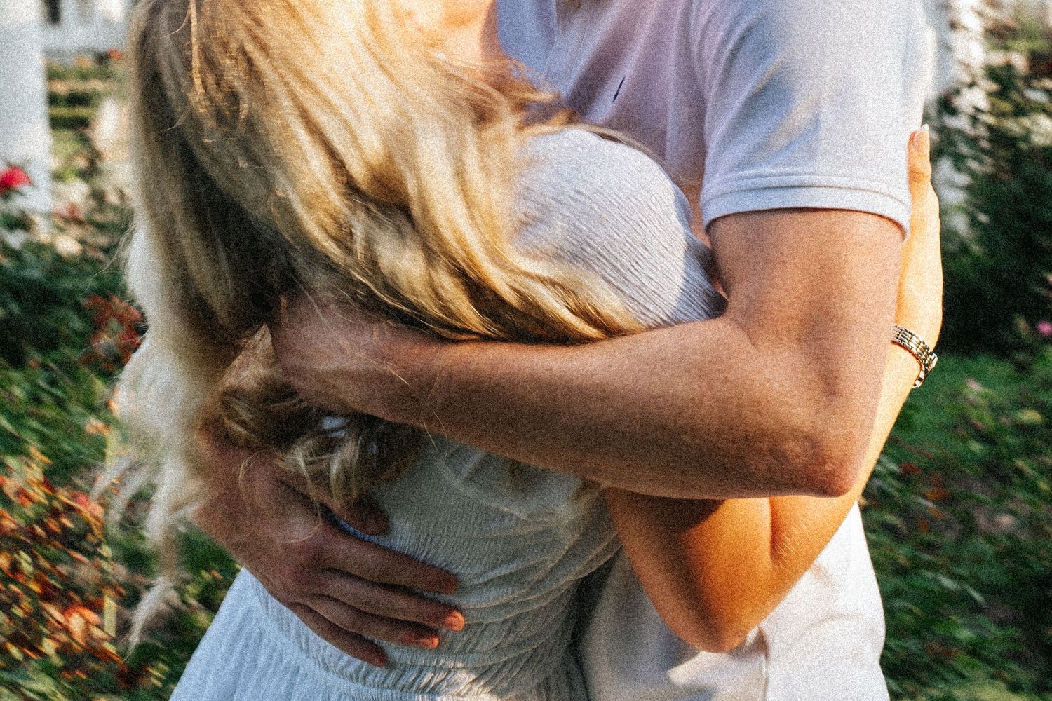 A couple stands together in an embrace against a green garden background in sunlight.