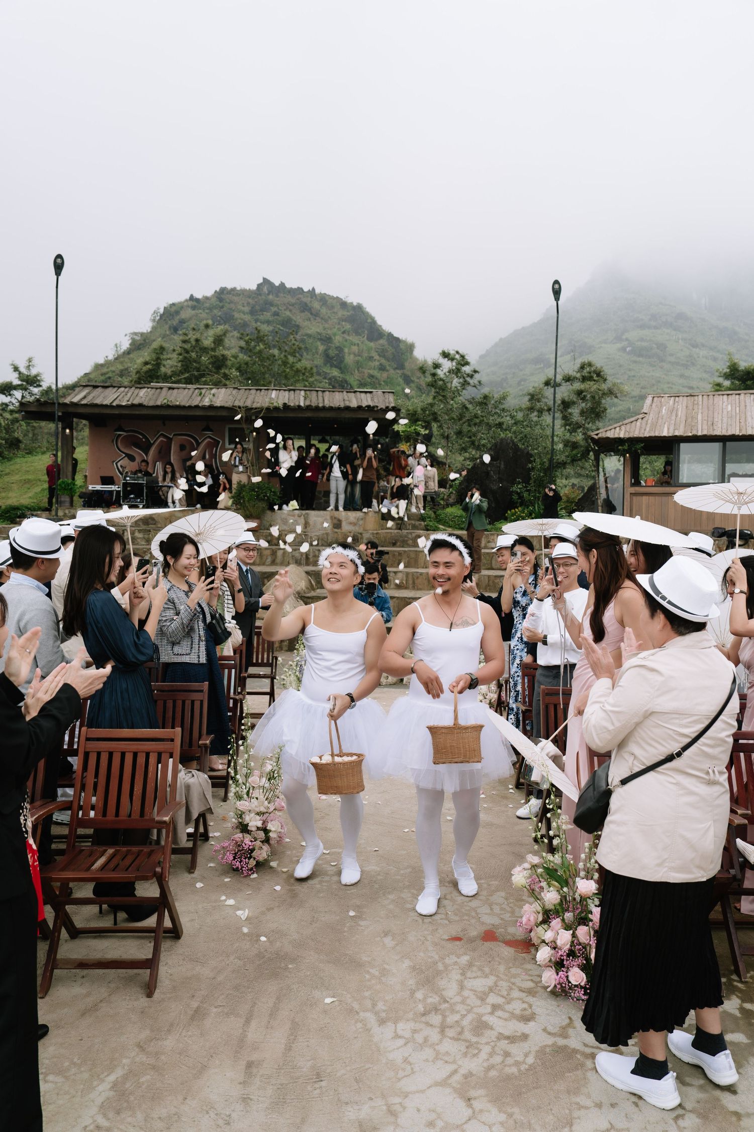 Two brides in white dresses and hats walk joyfully down the aisle as guests celebrate at an outdoor mountain wedding.