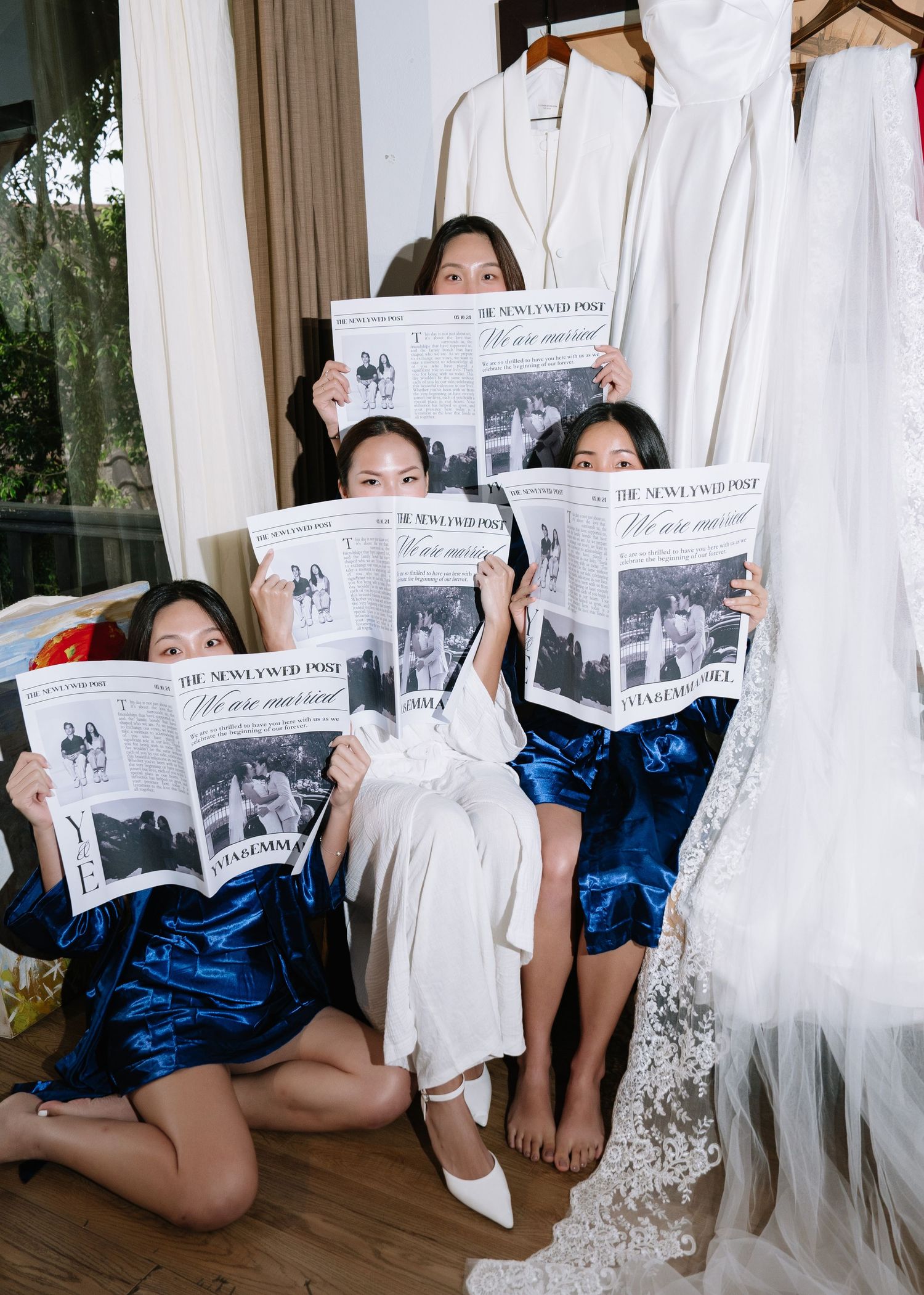 Bride and bridesmaids in robes holding wedding newspapers beside lace gown in bridal suite.