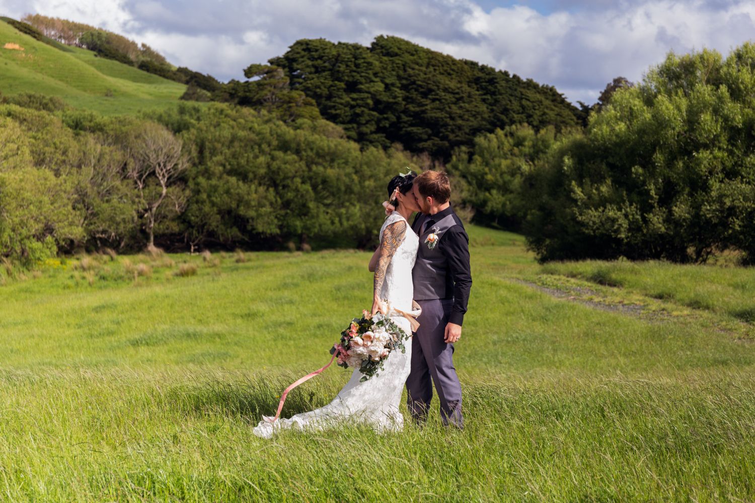 A couple shares a romantic moment in their wedding attire amid rolling green hills and lush coastal scenery.