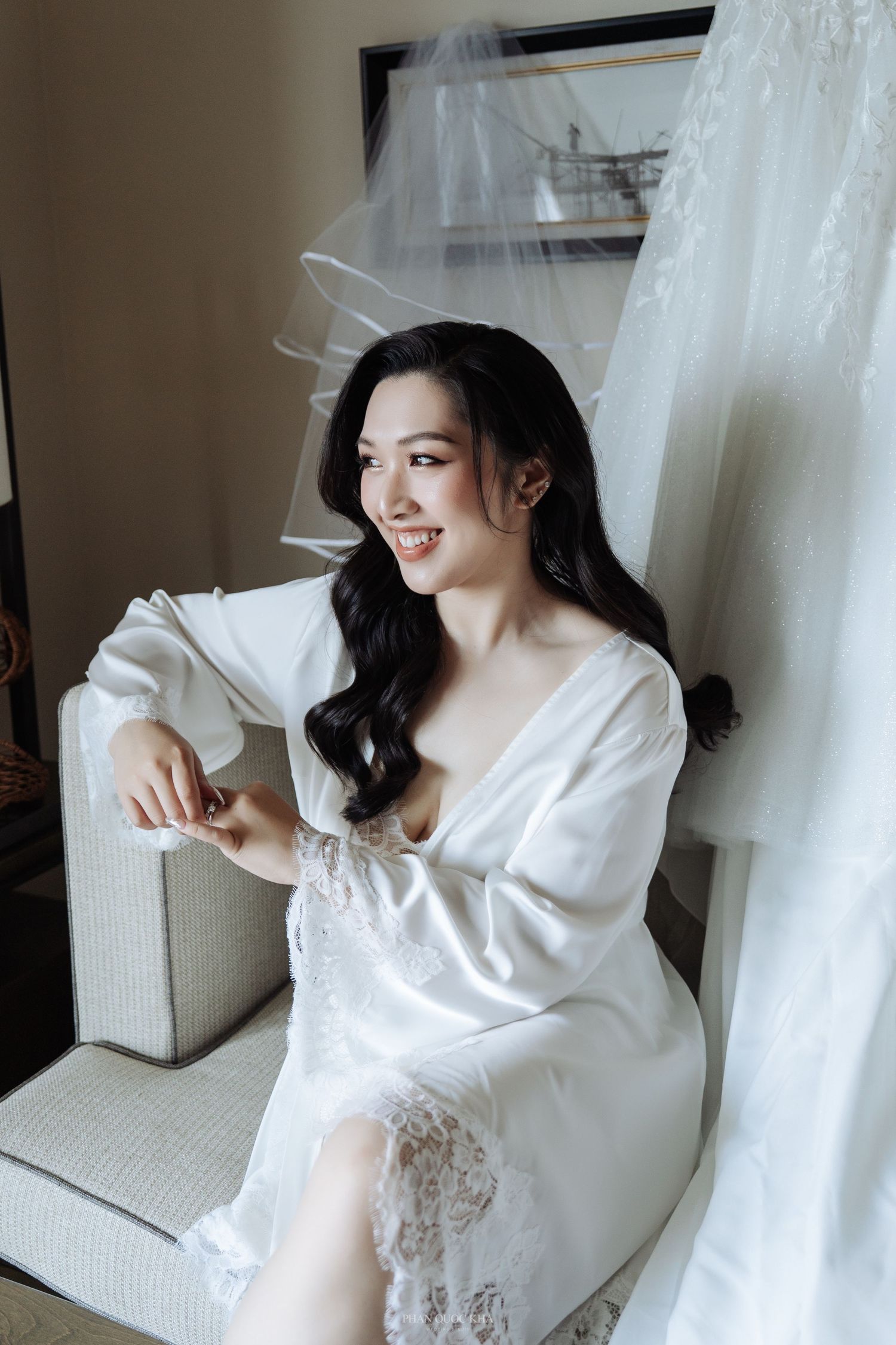 Beautiful smiling bride in a white lace robe sitting beside her wedding dress in a softly lit room.