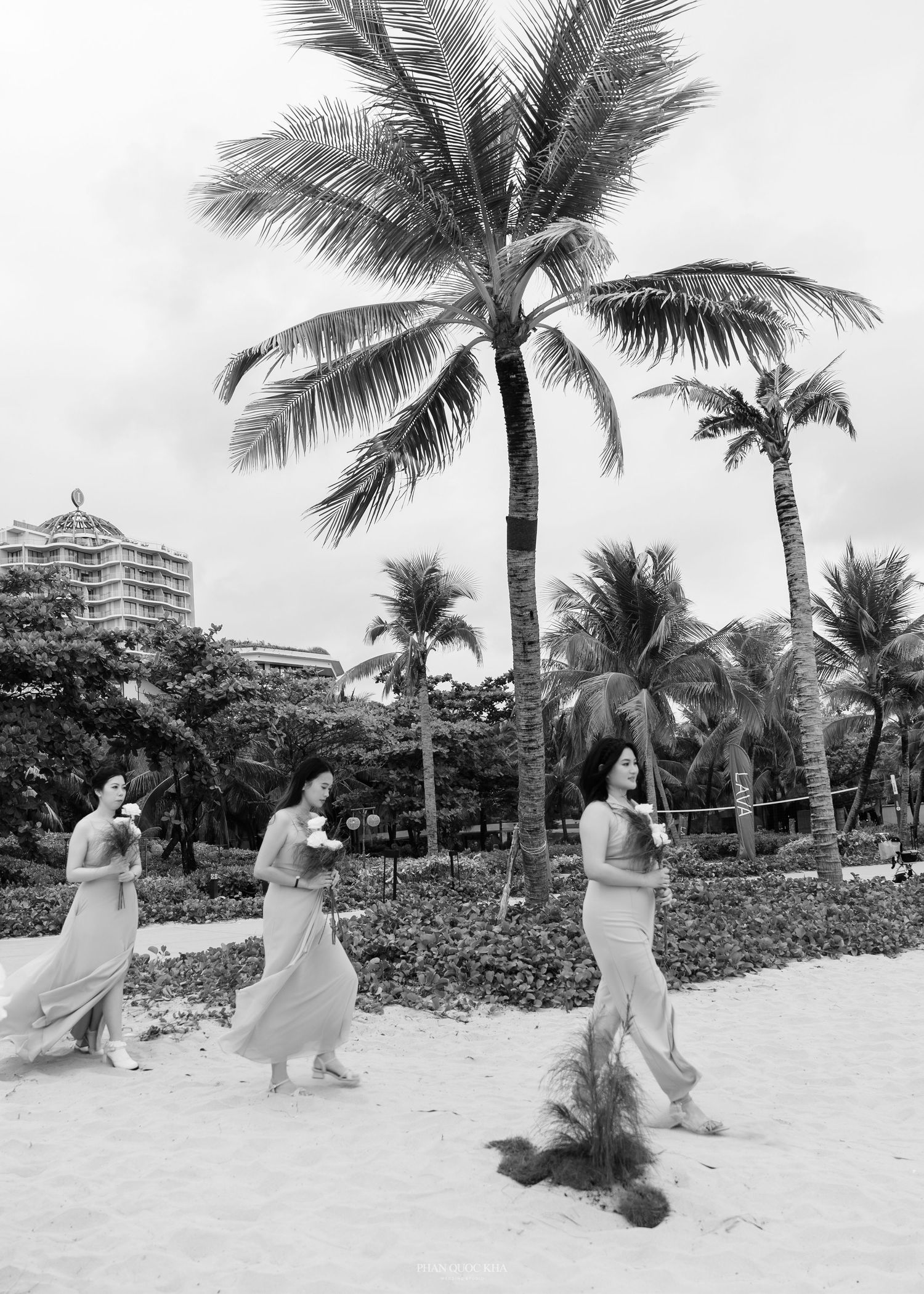 Black and white photo of three women in flowing dresses dancing on a tropical beach with palm trees.