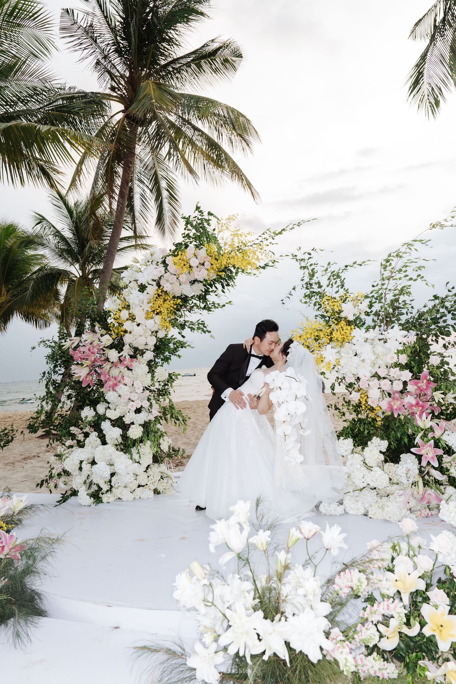 Bride and groom share a kiss under a floral arch adorned with white and yellow flowers on a beach.