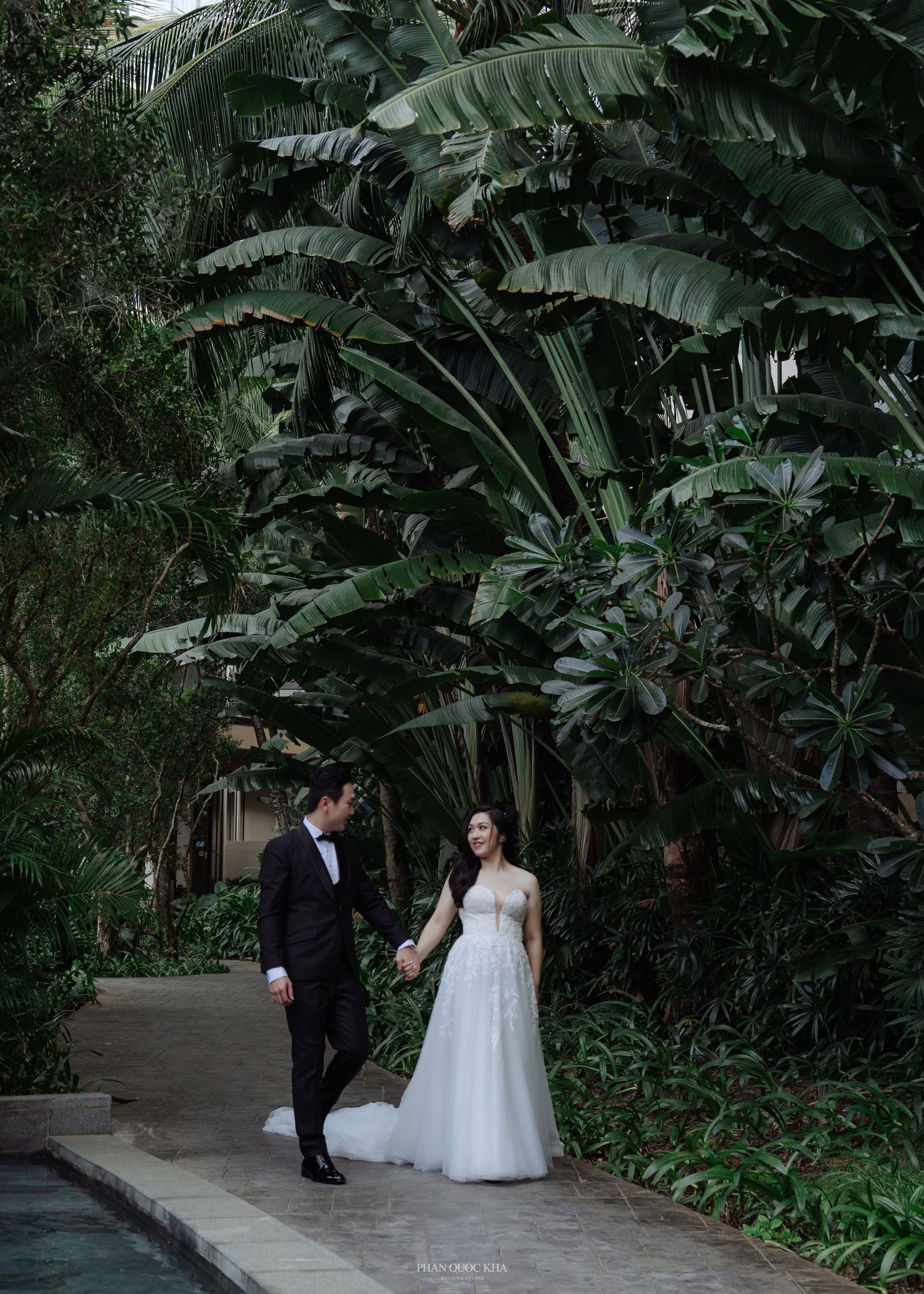 Bride and groom hold hands walking along a lush tropical garden path near a pool.