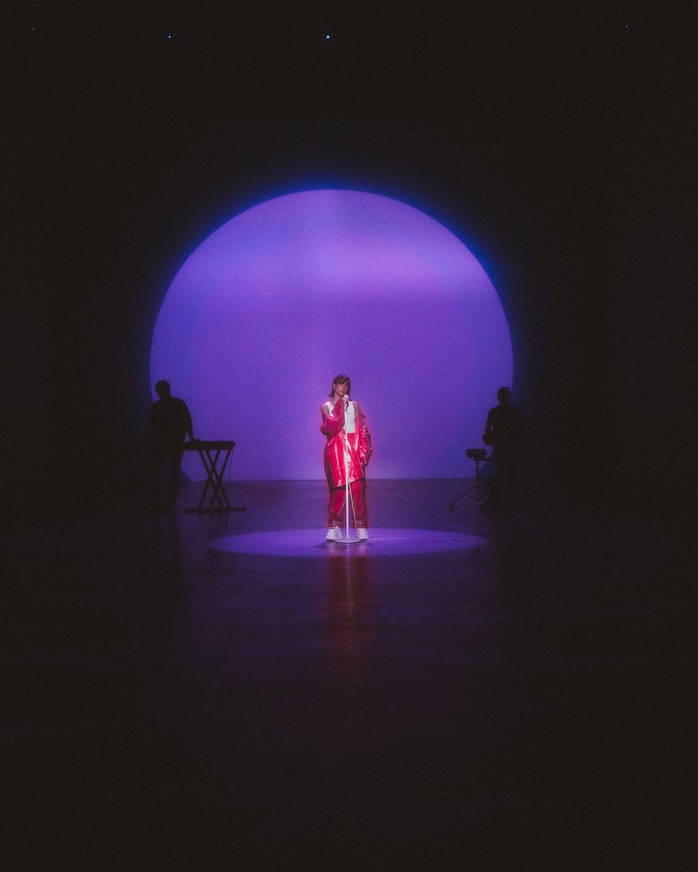 A performer in red attire stands in a spotlight on stage with a purple circular backdrop and silhouetted musicians.