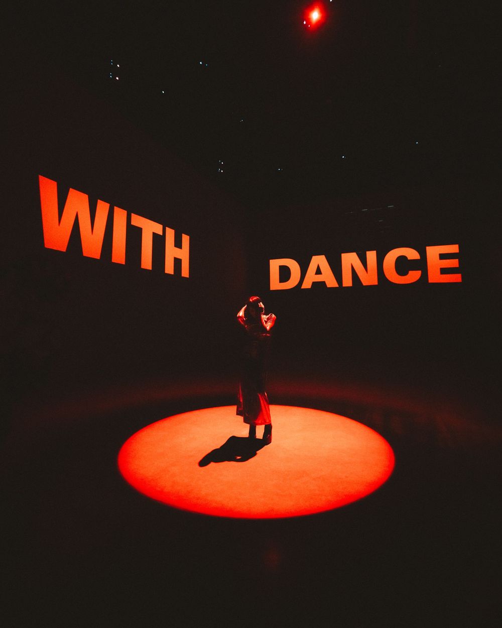 A silhouetted figure performs a dramatic dance move under a red spotlight on a dark stage.