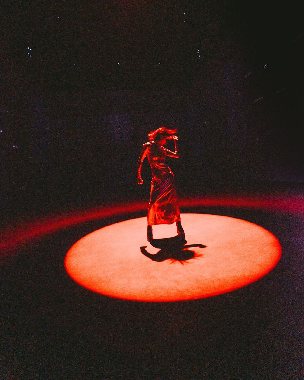 A dramatic silhouette of a dancer performing on a red-lit circular spotlight stage against a dark background.