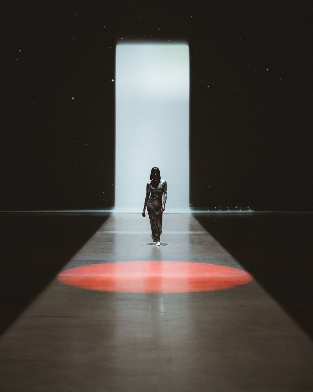Silhouetted figure walking on runway towards bright doorway with red circular spotlight on dark floor.