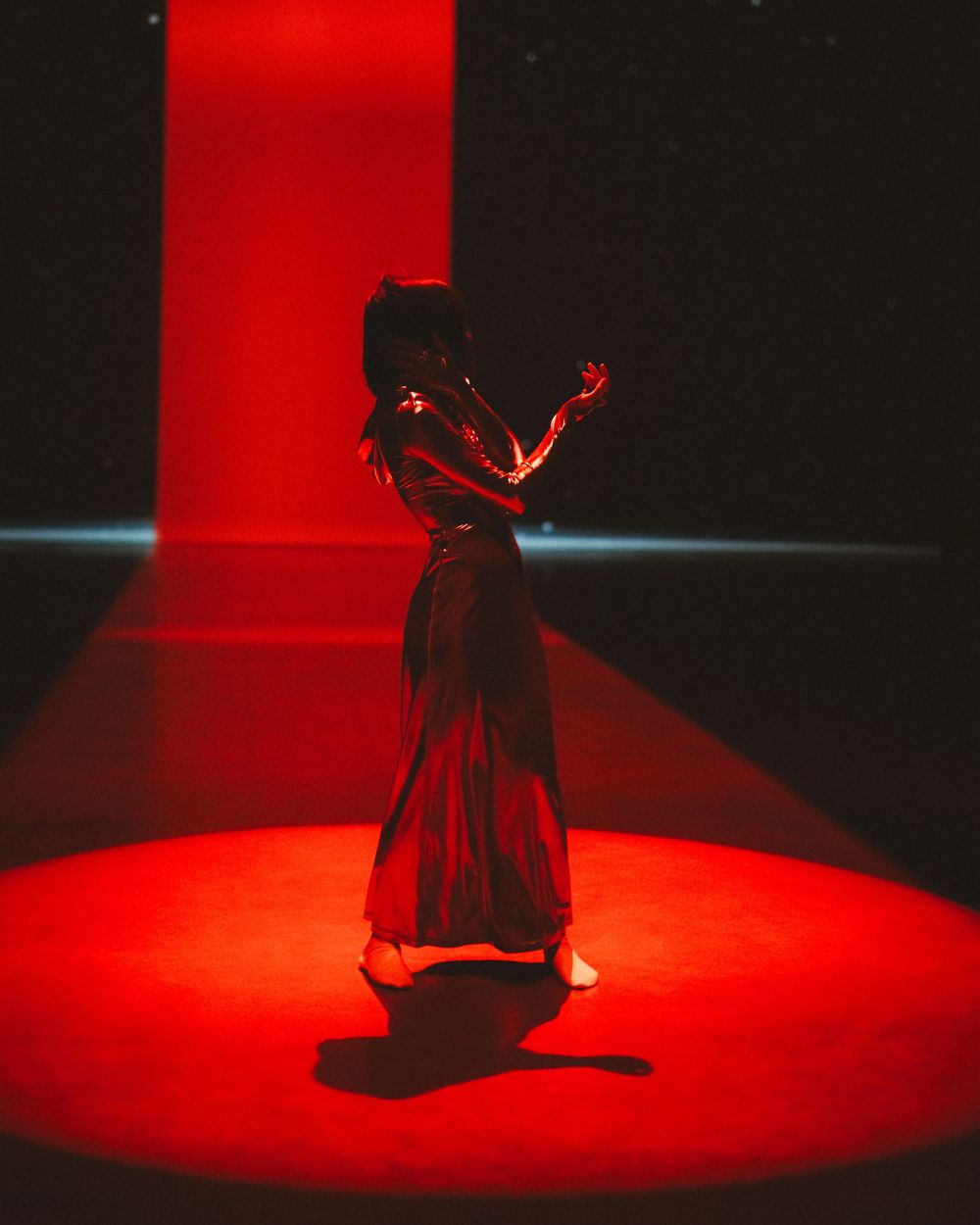 A silhouetted dancer performs in a dramatic red spotlight on a dark stage, casting a long shadow beneath.