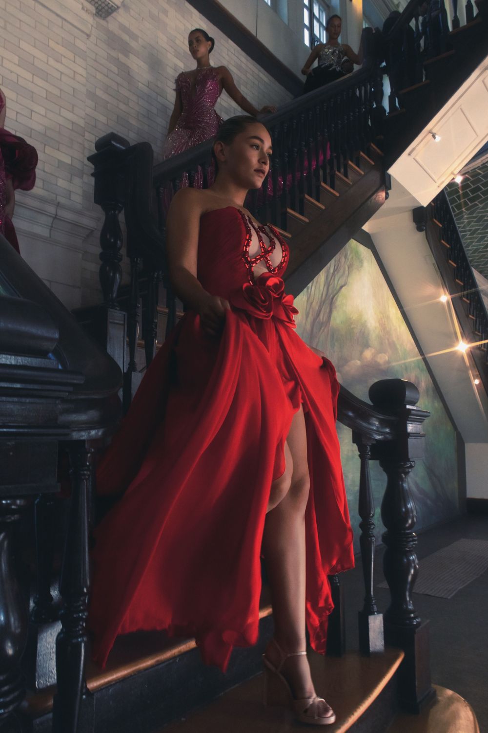 A figure in a flowing red dress poses on a staircase with dramatic lighting and shadows creating an elegant scene.