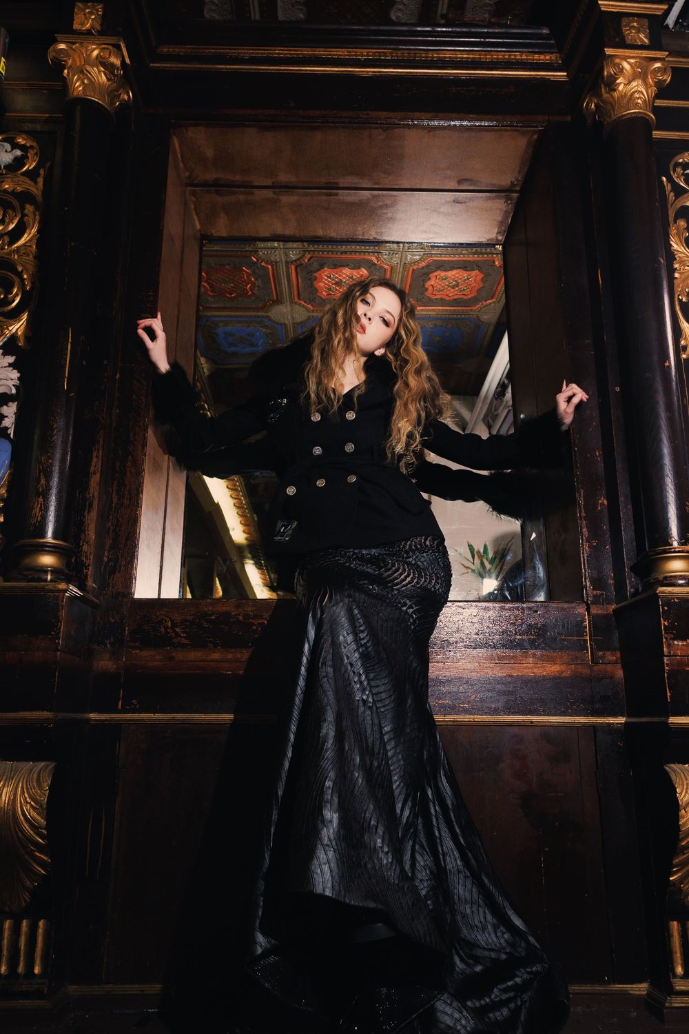 Fashion model poses in black evening gown against ornate dark wood interior setting.