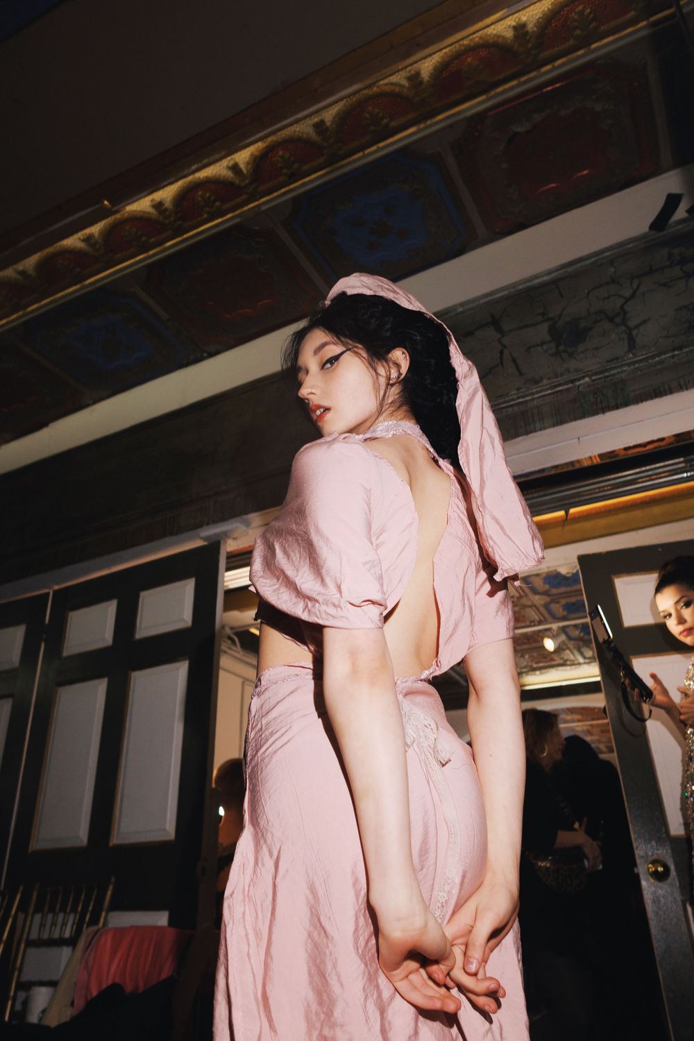 Model in pink two-piece outfit poses against dark industrial interior backdrop.