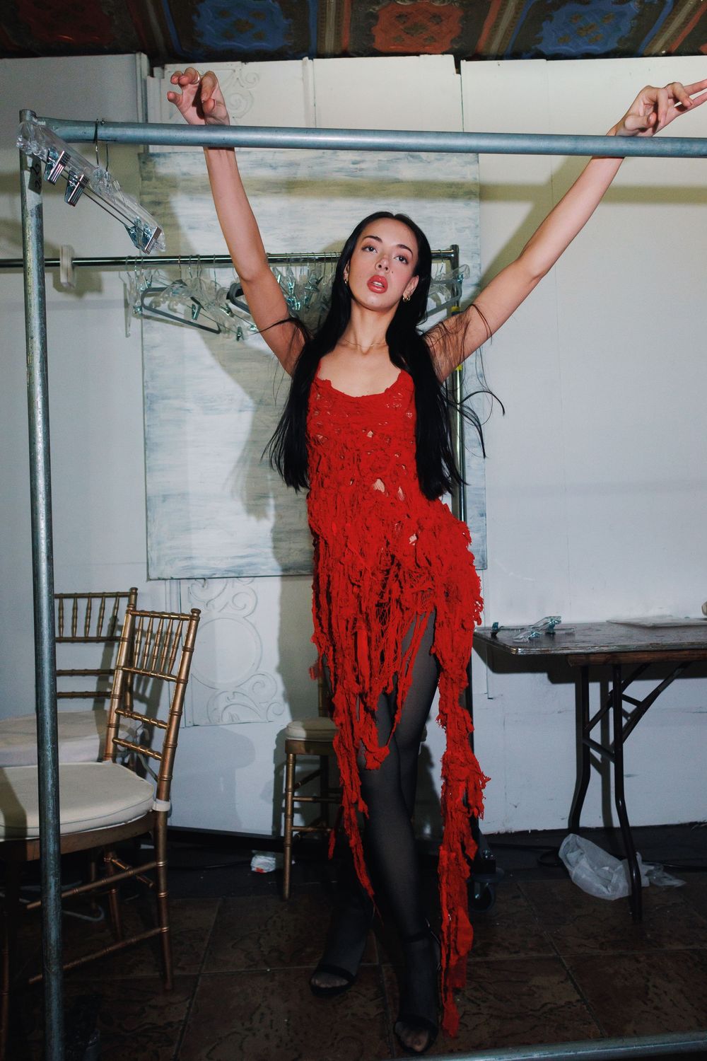 A person in a red fringed top and dark pants dances energetically in a room with wooden chairs against a white wall.