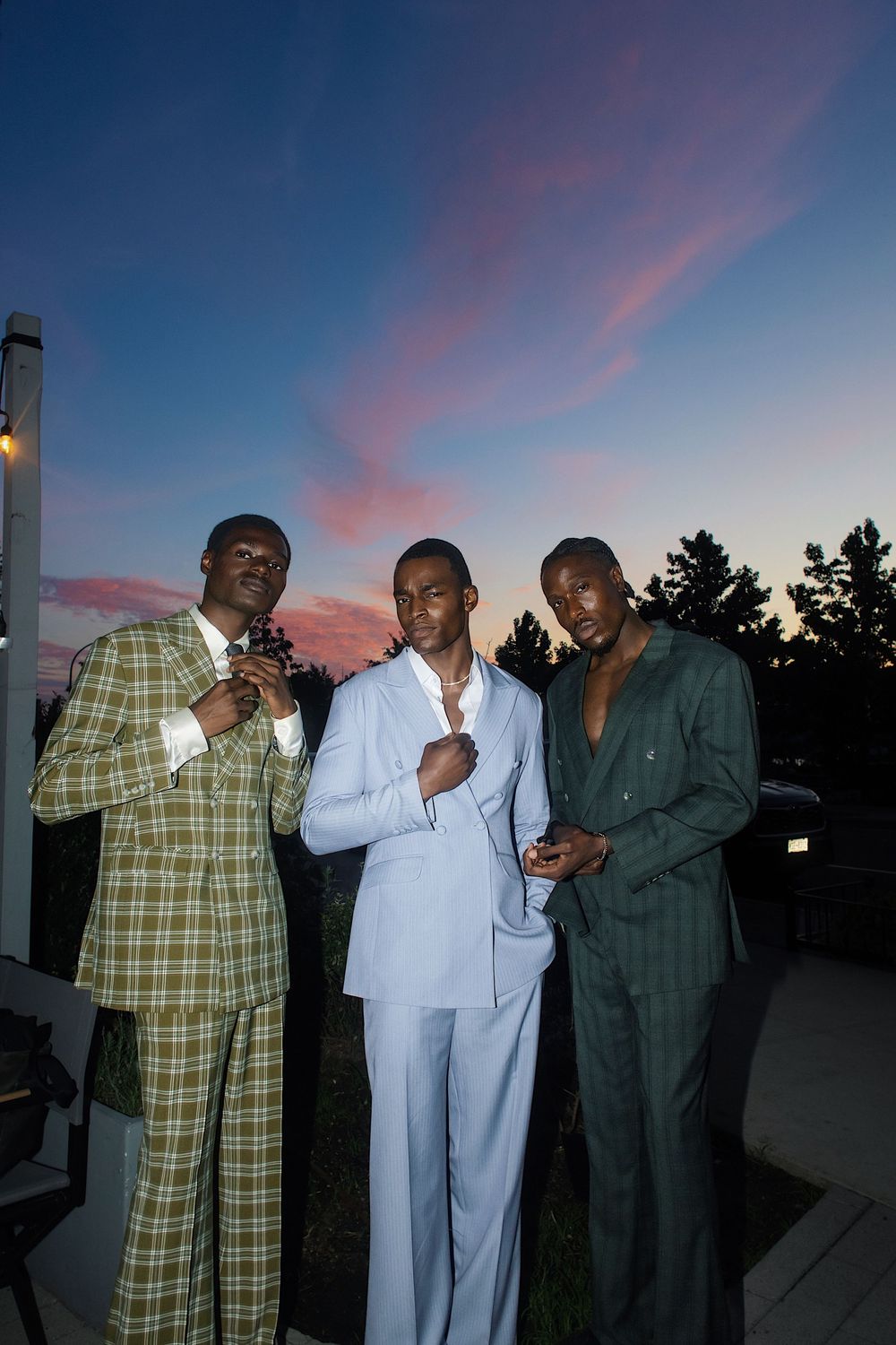 Three men in stylish suits pose together at dusk with pink clouds in the sky behind them.