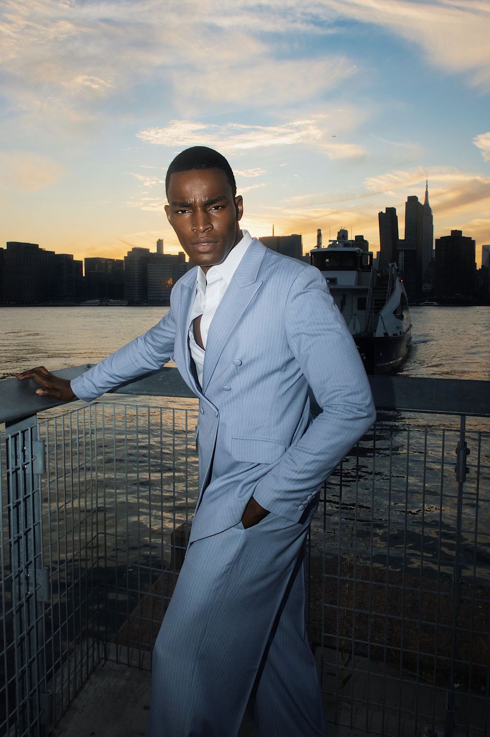 A person in a light blue suit poses against a sunset cityscape backdrop by the waterfront.